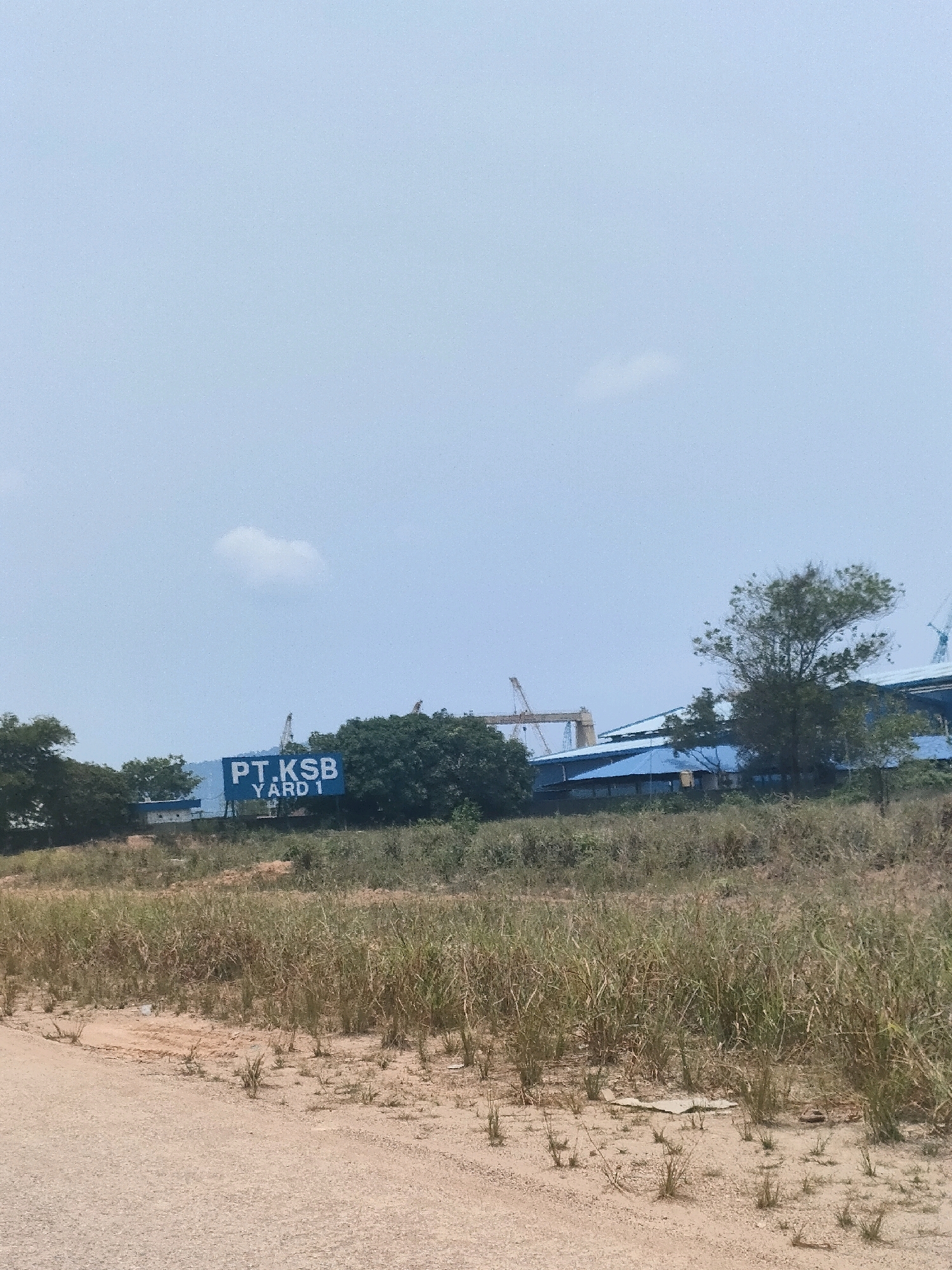 The image depicts a sign reading "PT. KSB YARD 1" set against a backdrop of industrial buildings and trees under a pale, hazy sky. The sign itself is blue with white lettering and appears to be mounted on a pole or structure. To the left of the sign, there are a few trees, and behind it, a large blue-roofed building with some visible cranes and industrial structures. The foreground consists of dry, patchy vegetation and a dirt path. The scene suggests an outdoor industrial area, likely a shipyard or construction site, located in Batam, Indonesia. There are no people visible in the image, and the overall impression is one of a functional, operational facility.