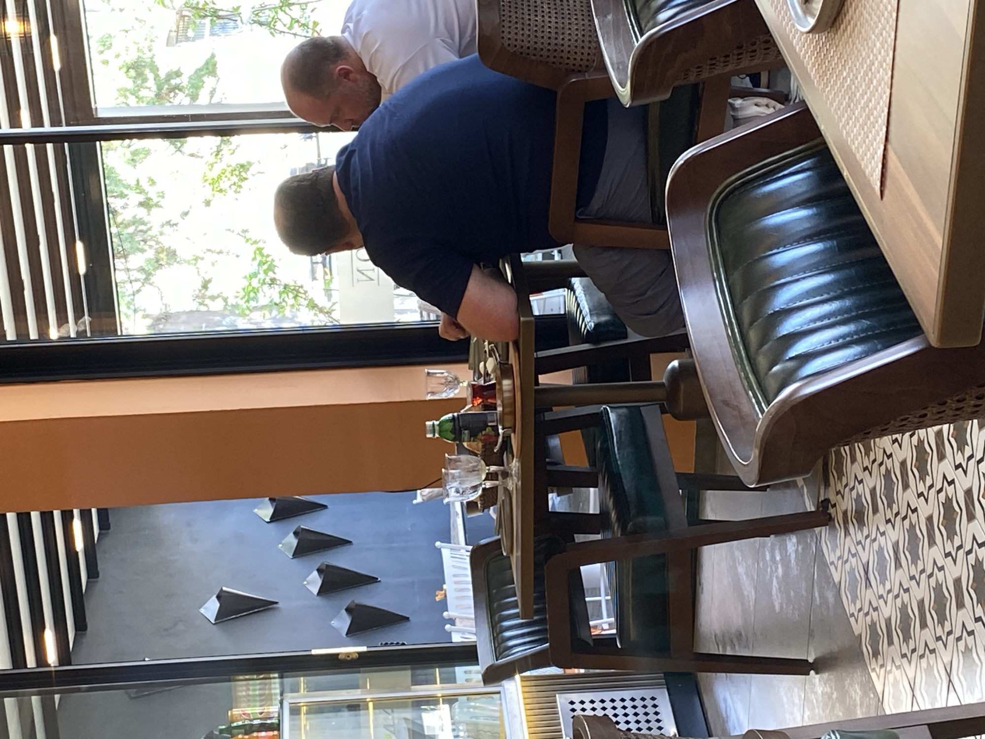 The image depicts the interior of a restaurant with a view of trees and foliage through large windows. Two men are seated at a table, their backs to the camera. The man on the left wears a dark blue t-shirt and grey pants. The man on the right wears a white t-shirt. A partially visible table is set with glassware, a bottle, and some food items. The restaurant features dark-colored chairs with green upholstered seats and wooden frames, and a patterned tile floor. On the wall in the background, several dark, cone-shaped decorative items are mounted. The scene appears to be in Sokhumi, Abkhazia.