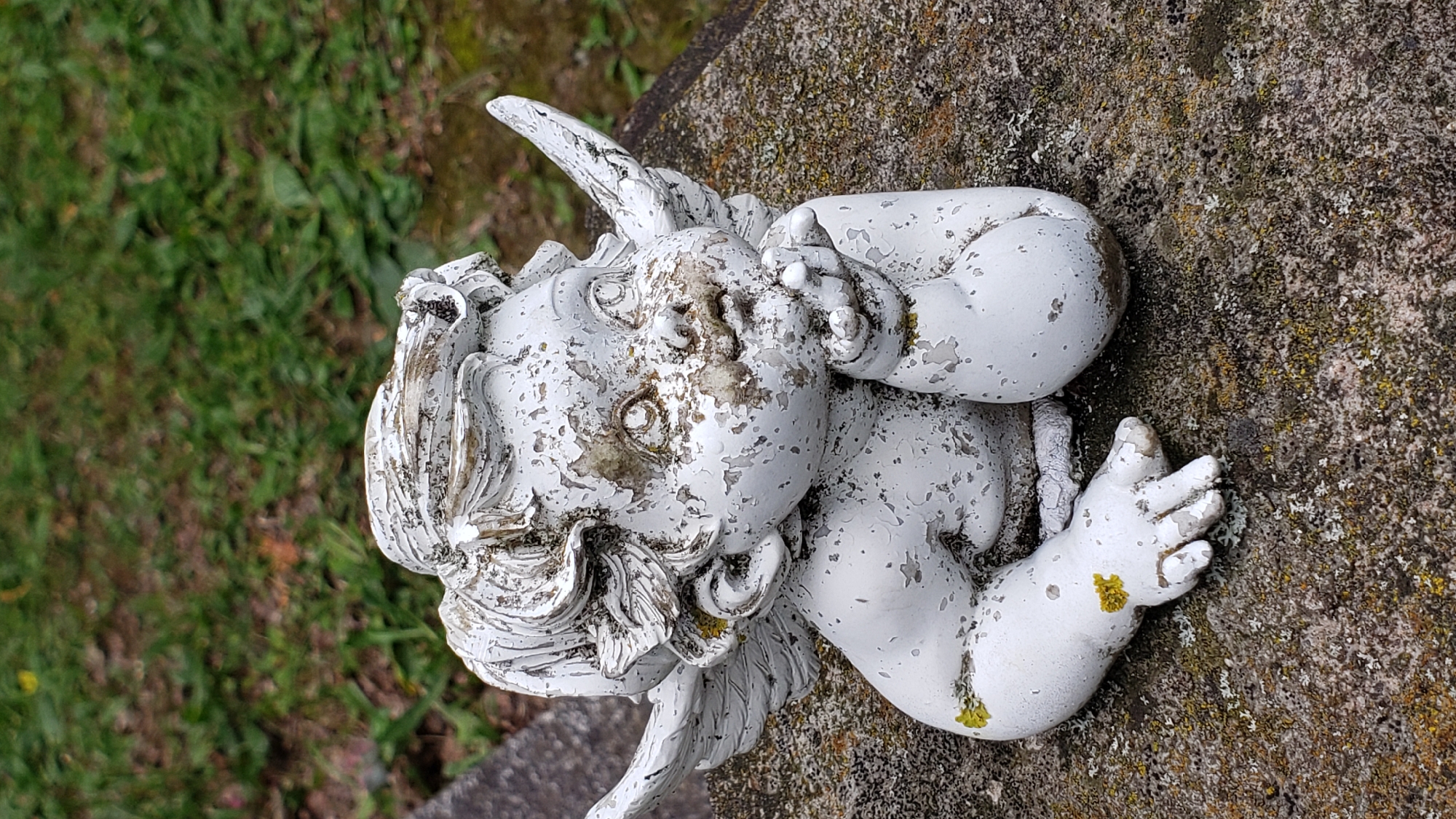 An aged, white cherub statue, exhibiting significant chipping and weathering with patches of green and brown lichen, rests on a rough-textured, grey stone surface. The statue is positioned with its left side towards the viewer, leaning on its left arm and propping its head with its right hand near its chin. Its wings are partially visible. The background consists of out-of-focus green grass and foliage. This scene is located on a tombstone in the public cemetery of Prešov, Slovakia.