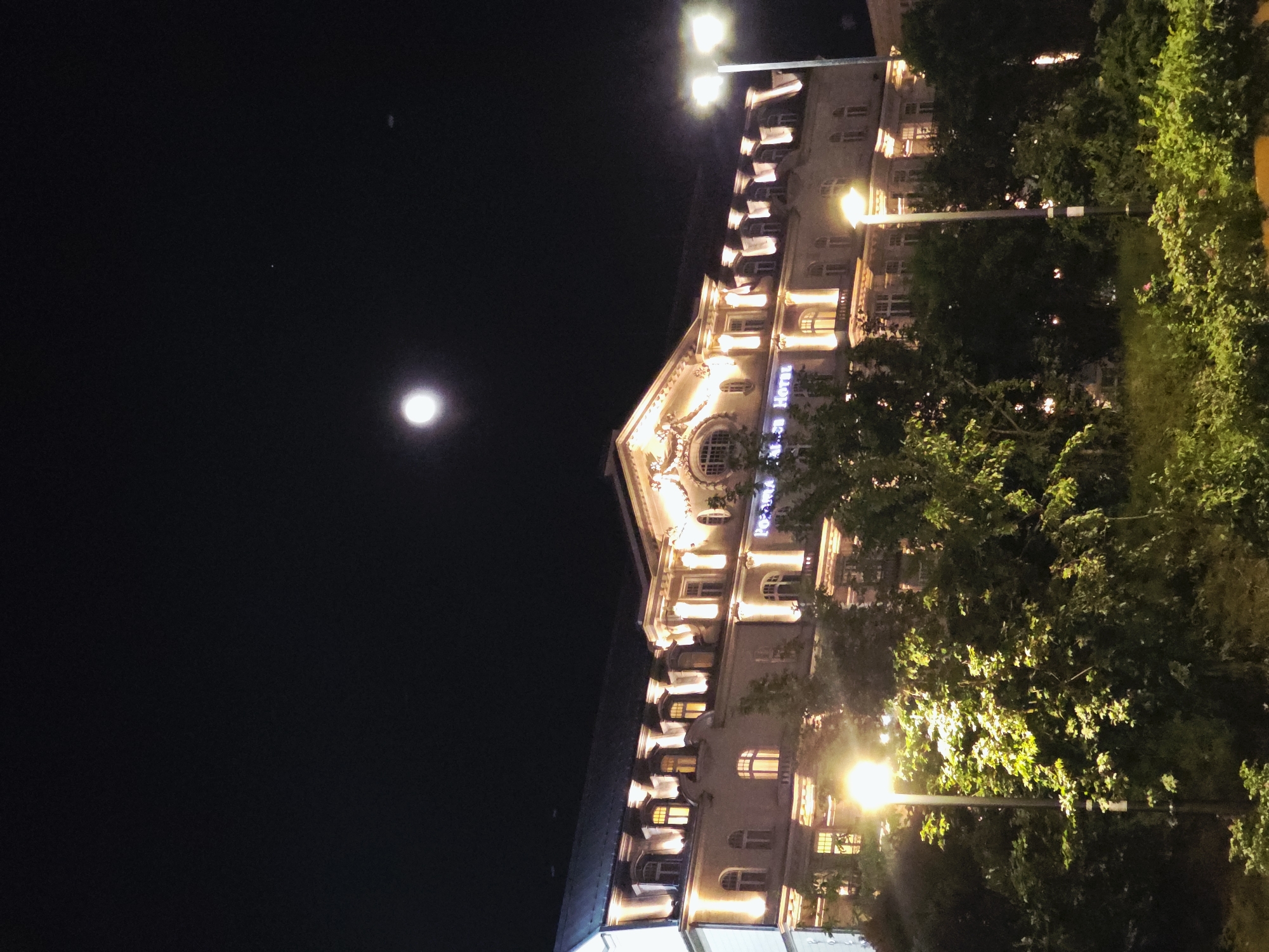 The photograph depicts a nighttime scene in Warsaw, Poland, dominated by a multi-story building illuminated by artificial lights. The building's facade exhibits architectural details and includes a sign that appears to read "Hotel" in Polish. The sky is dark, with a full moon visible in the upper left corner. A row of trees flanks the right side of the building, adding a natural element to the urban landscape. Streetlights cast a glow, highlighting both the building's architecture and the surrounding vegetation.