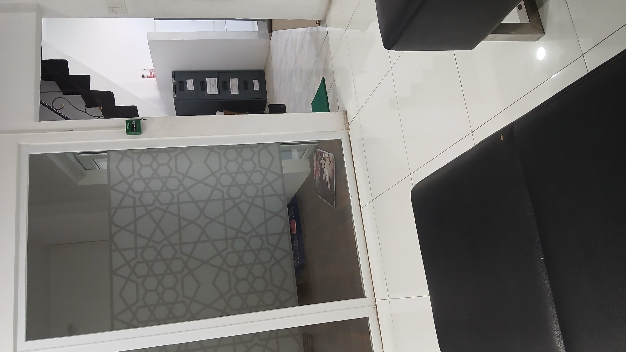 The image shows an interior space, possibly an office or waiting area, in Lhokseumawe, Indonesia. The space is dominated by light-colored tile flooring and white walls. There is a set of black stairs with decorative railing visible in the upper left corner, leading to an unseen upper level. Near the stairs are filing cabinets. There's a frosted glass window with a geometric pattern. Two black chairs are visible in the foreground, suggesting a seating area. The orientation suggests the image was taken from a slightly elevated perspective.