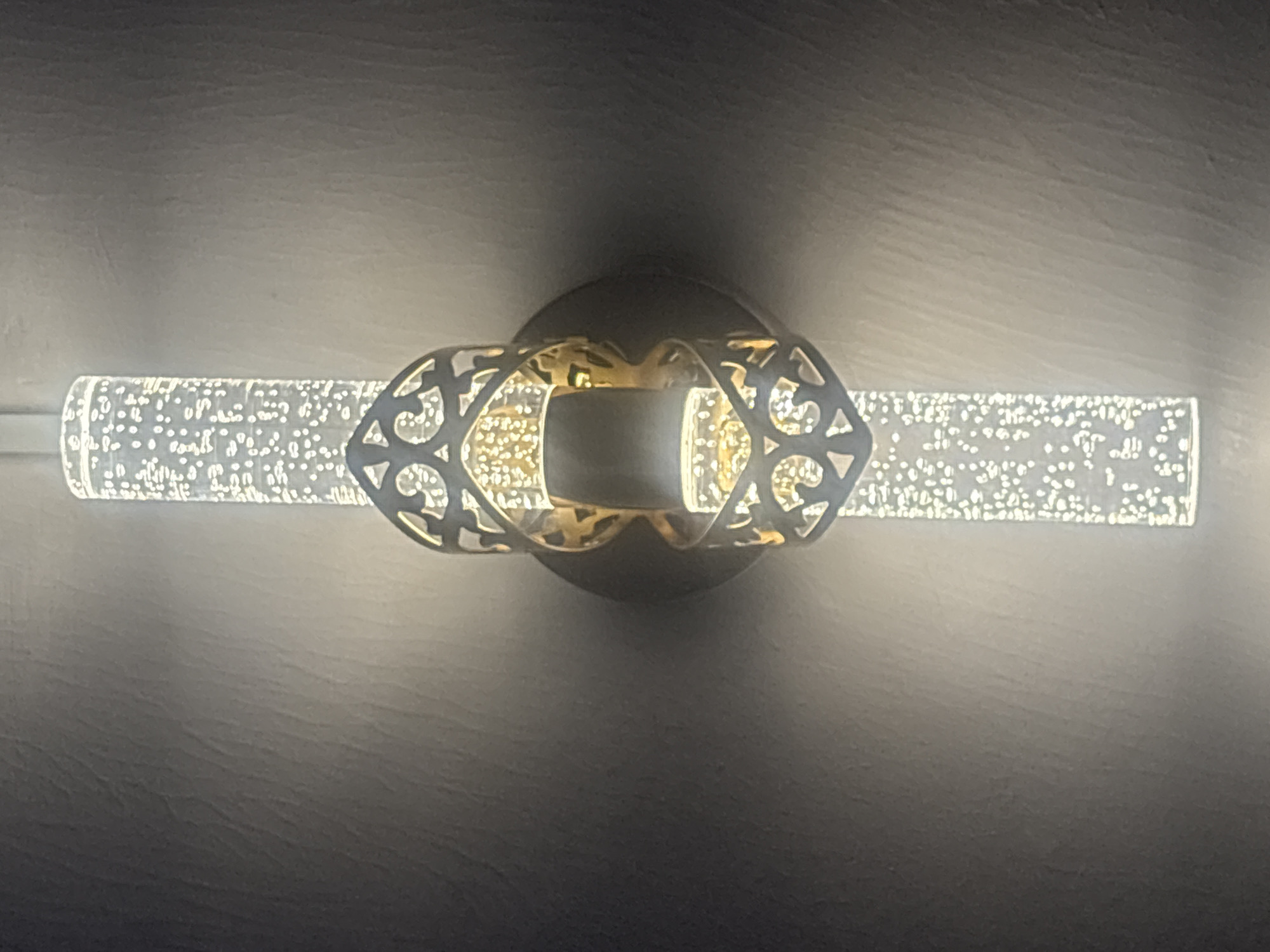 The image shows a light fixture mounted on a wall. The fixture consists of a central, round base from which a cylindrical light extends horizontally on either side. The cylindrical lights are filled with small, reflective particles that scatter the light. Decorative, metallic filigree surrounds the central section where the lights attach to the base. The wall appears to have a slightly textured surface. There is no information available to infer the location is Rayfield, Nigeria.