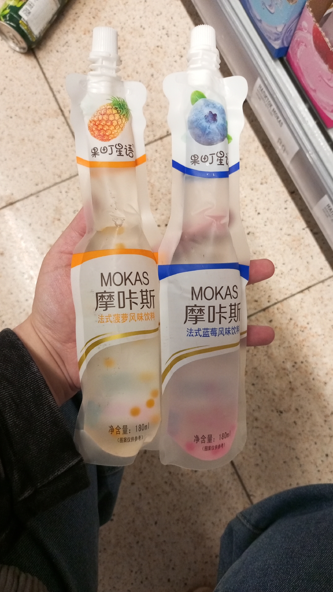 The image shows a hand holding two squeezable pouches of MOKAS beverages. The pouch on the left features a pineapple illustration and is labeled as "French pineapple flavor drink". The pouch on the right features a blueberry illustration and is labeled as "French blueberry flavor drink". The beverages have small colored balls suspended within. The net content is marked as 180ml for both. The location appears to be a store, as indicated by the shelving visible in the background. The context of Трубачеевка, Russia, is not directly evident from the image.