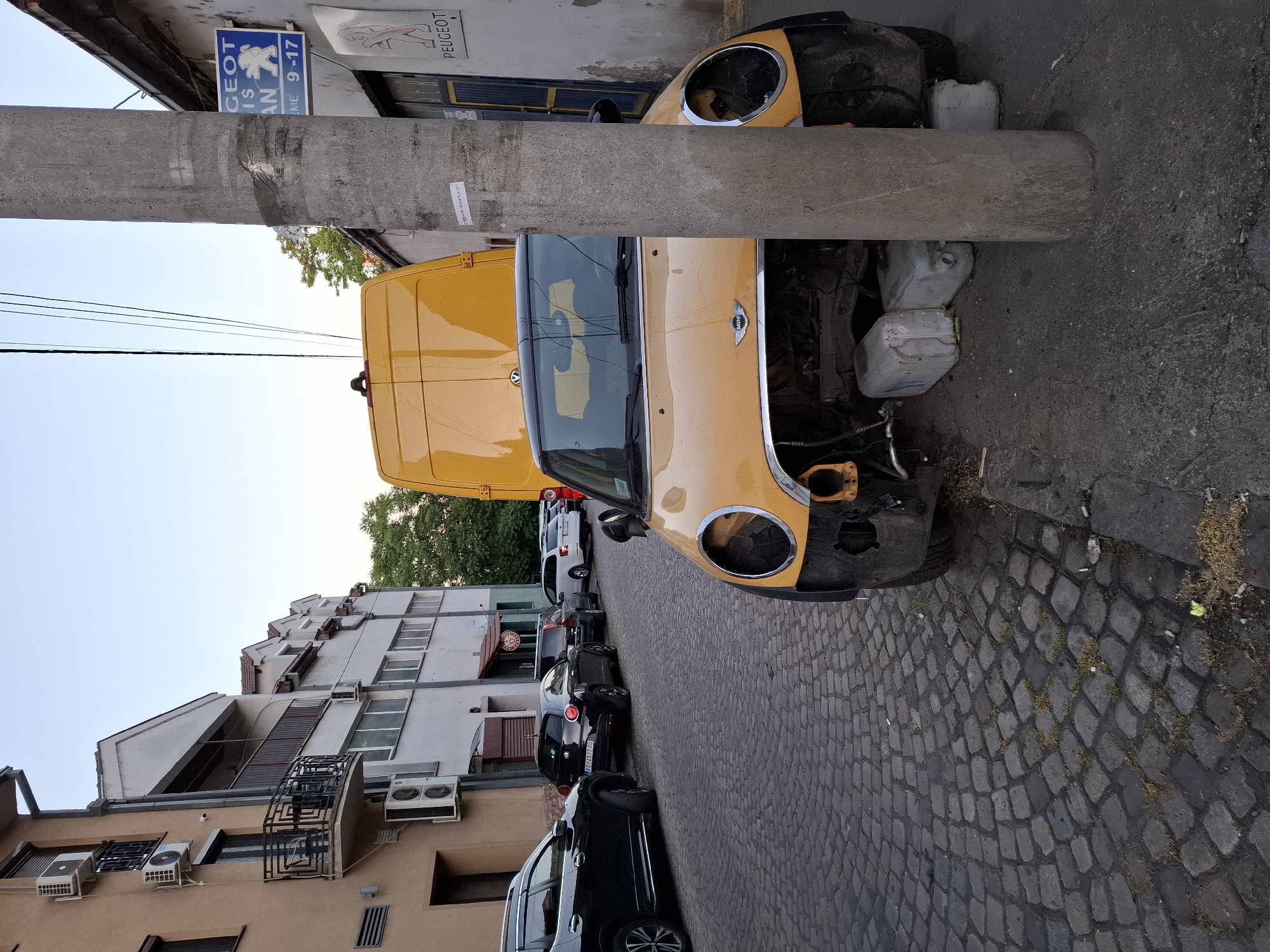 The image shows a street scene in Zemun Urban Municipality, Serbia. A yellow van and a yellow Mini Cooper without headlights are parked next to a concrete pole on a cobblestone street. Other parked cars are visible in the background, along with buildings featuring air conditioning units and a fire escape. A sign indicates "GEOT IS AN" and hours "ME 9-17". Empty plastic containers are visible next to the Mini Cooper. No people are clearly visible in the frame, although a person is standing in the open van.