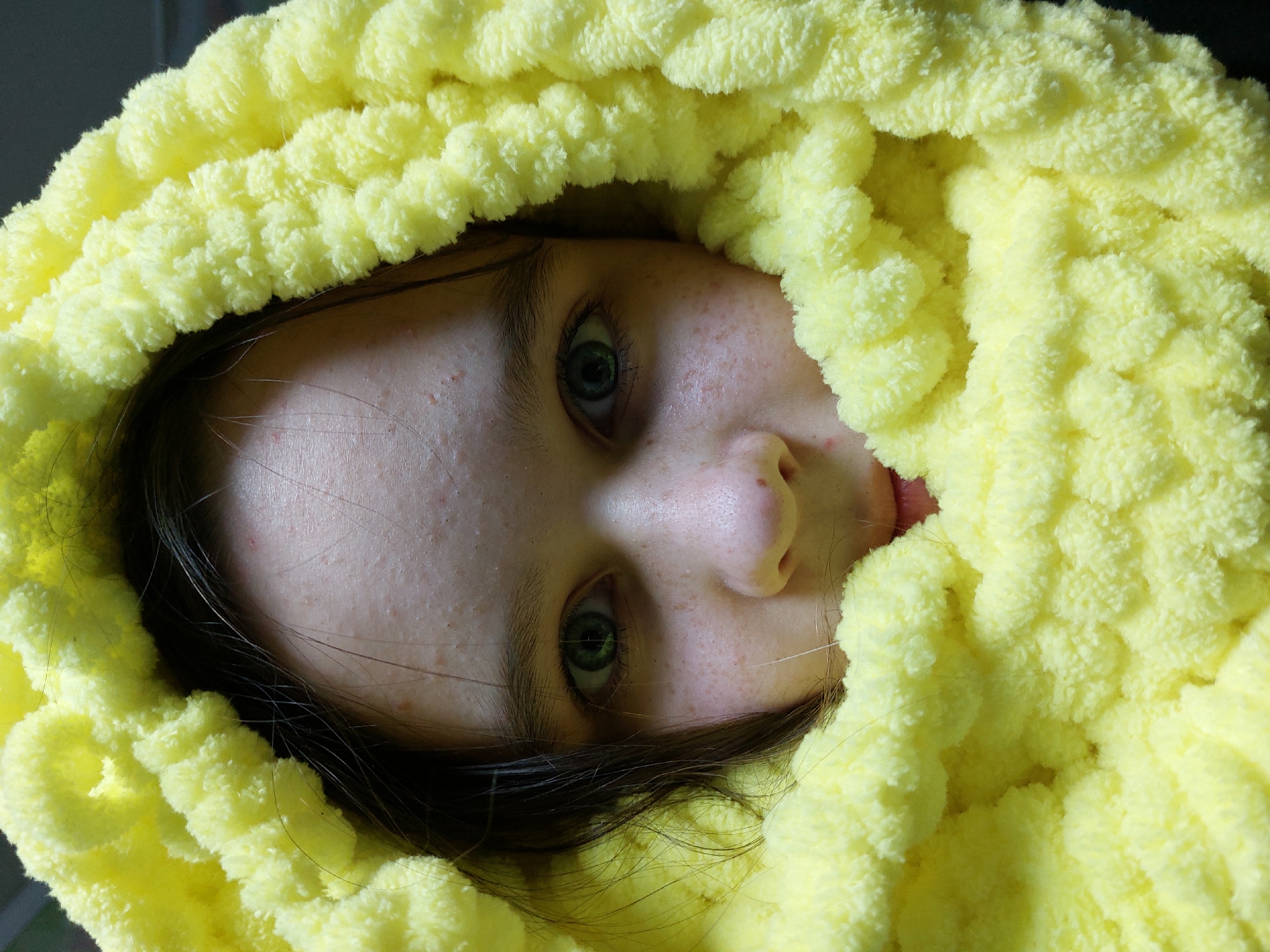 A close-up, high-angle portrait captures a young individual's face peeking out from a vibrant, fluffy yellow material, likely a blanket or large scarf. The image is rotated 90 degrees clockwise, orienting the person's head with their left side facing upwards, and their gaze directed towards the viewer. The subject's most striking features are their clear blue-green eyes. Their fair skin shows scattered freckles or minor blemishes, and dark strands of hair are visible around the face.
The dominant object in the scene is the bright yellow material, which is thick, chunky, and has a soft, plush texture, characteristic of chenille or similar looped yarn. It frames the face almost entirely, creating a sense of coziness or being nestled. The background is dark and out of focus, drawing all attention to the face and the yellow fabric. No specific activity is depicted other than the person being covered and looking out. The setting appears to be indoors, with no further location cues visible within the image itself. The photograph's context is Chaikovsky, Russia. The time of day is not discernible, and the expression on the person's face is neutral, conveying a calm or curious demeanor. There is no visible text in the image.