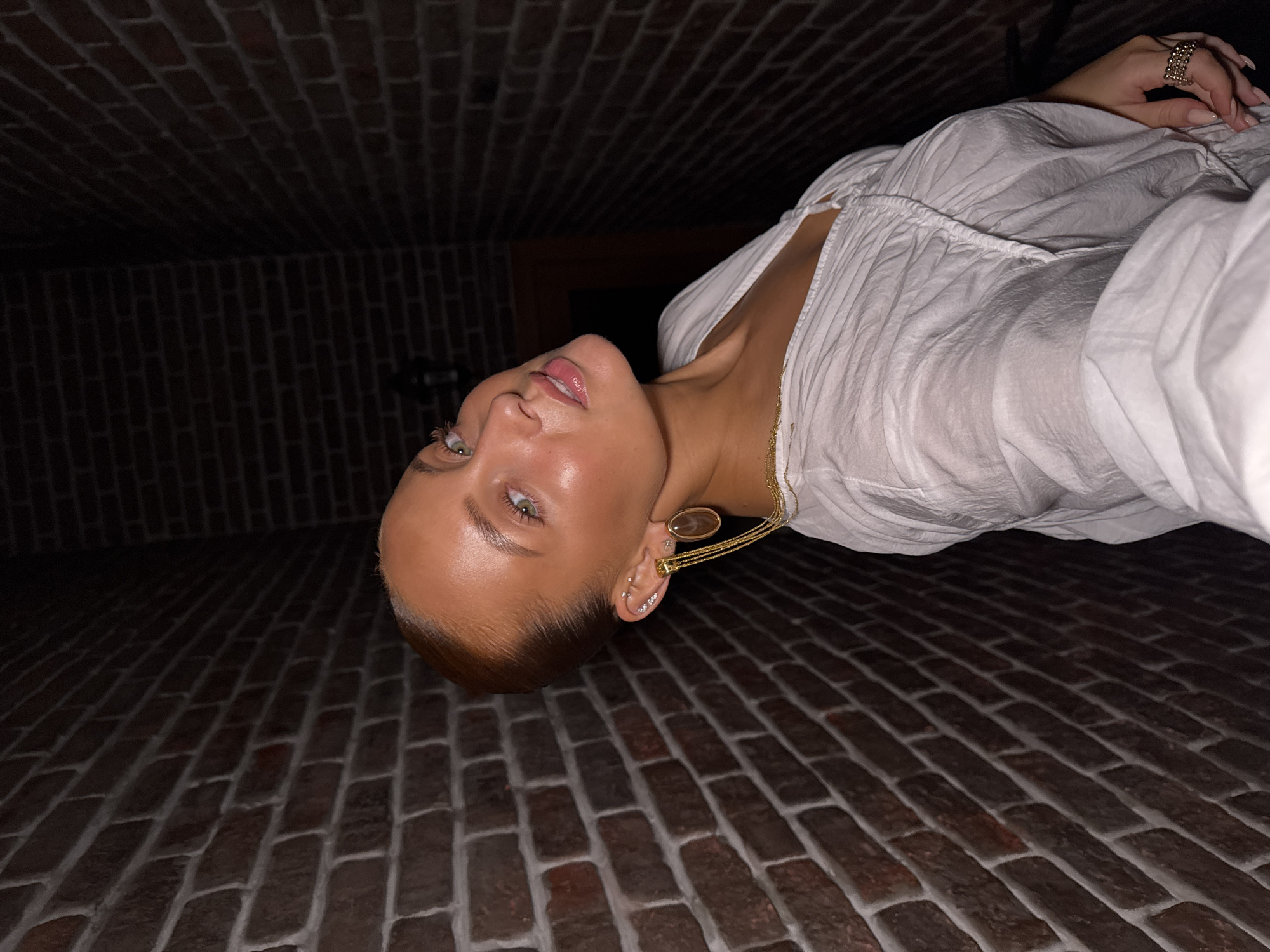 A young woman with auburn hair pulled back is looking towards the camera. She is wearing a white ruffled shirt and a gold earring with an oval-shaped pendant. Her makeup is highlighted with shimmer on her cheeks and lips. The background is a dark brick wall, which appears to be located in a dimly lit indoor setting. There are no discernible elements in the image that would indicate the specific location within Chaikovsky, Russia.