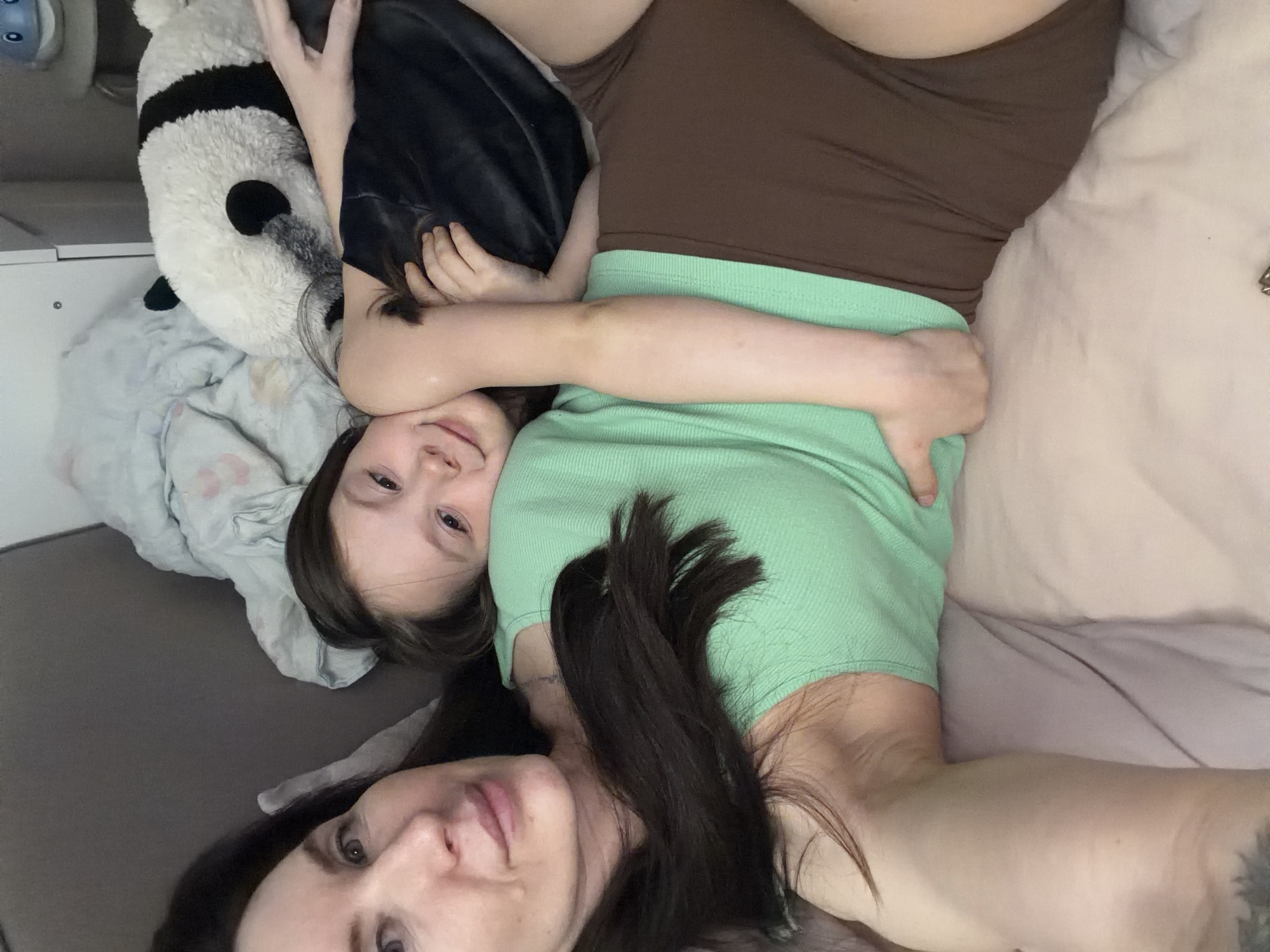 Two individuals, an adult female and a child, are captured in a close-up, overhead shot while lying down indoors, possibly in a bedroom in Perm, Russia. The image is rotated, presenting the subjects as if they are lying on a vertical plane.
In the foreground, the adult female is visible, showing her face with dark hair, a light green ribbed top, and brown shorts or leggings. Her right arm is gently wrapped around the child's upper body. She has a neutral to slightly serene expression with her lips slightly parted.
The child is positioned next to the adult, resting their head near the adult's shoulder. The child's face, also with dark hair, is visible, displaying a soft, gentle expression. A large, black and white panda plush toy is nestled beside the child, and the child's arm is draped over it.
The setting appears to be a comfortable indoor space. To the right, a light-colored sheet or duvet is visible. On the left, a grey blanket or surface is present, with a lighter grey patterned blanket (possibly with stars or dots) beneath the child and the panda. A white piece of furniture, possibly a bedside table, can be seen in the upper left background.
The overall impression is one of affection, comfort, and relaxation between the two individuals. There is no visible text in the image.