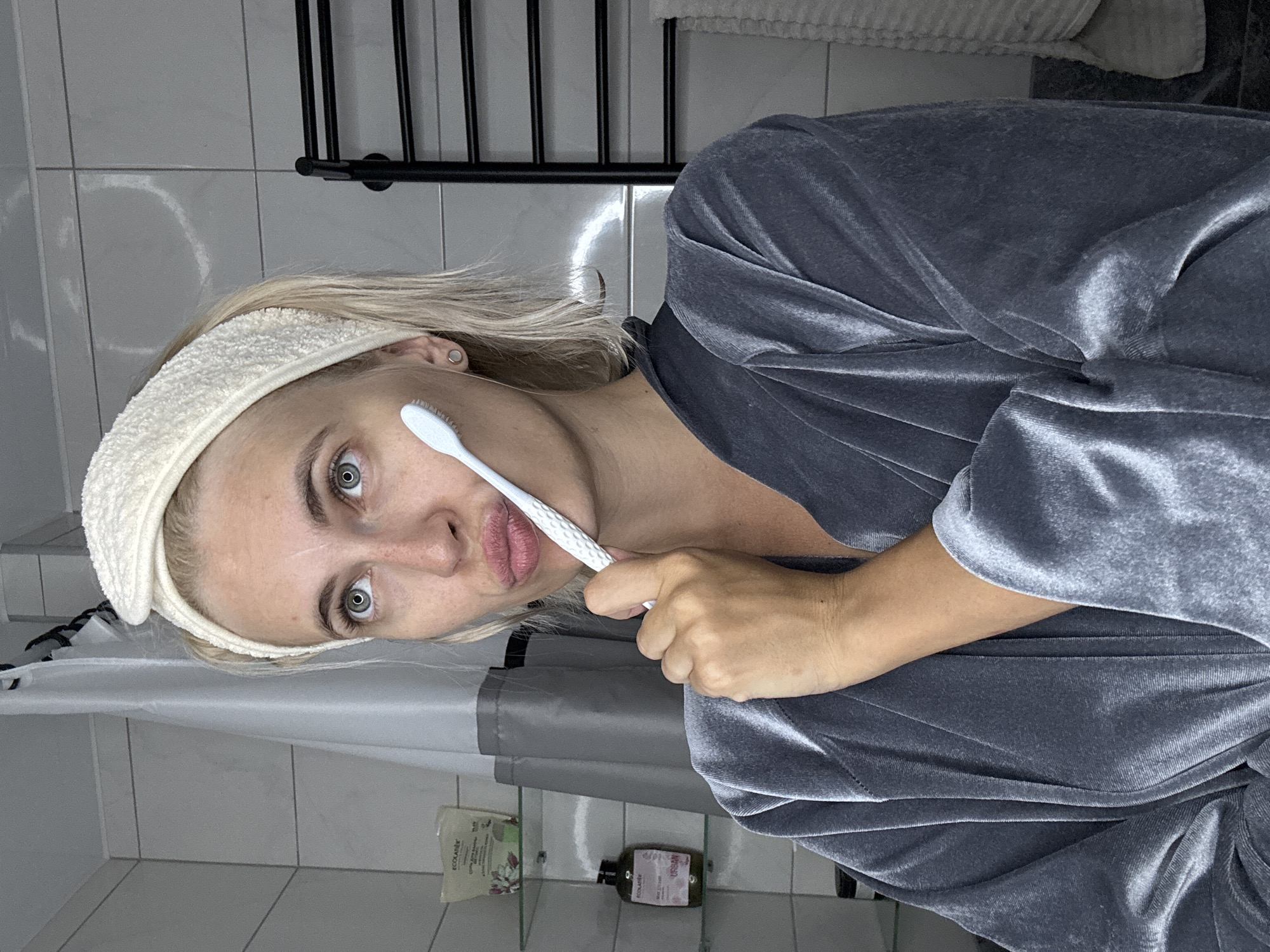 A young Caucasian woman with blonde hair, wearing a white terrycloth headband and a grey velvet robe, holds a white facial cleansing brush to her cheek while looking upwards. She is in a bathroom with white tiled walls, a grey shower curtain, and shelves containing beauty products. To the right, a black towel rack is visible. The scene appears to be indoors, likely in a domestic setting. The location of Saratov, Russia is not discernible from the image.