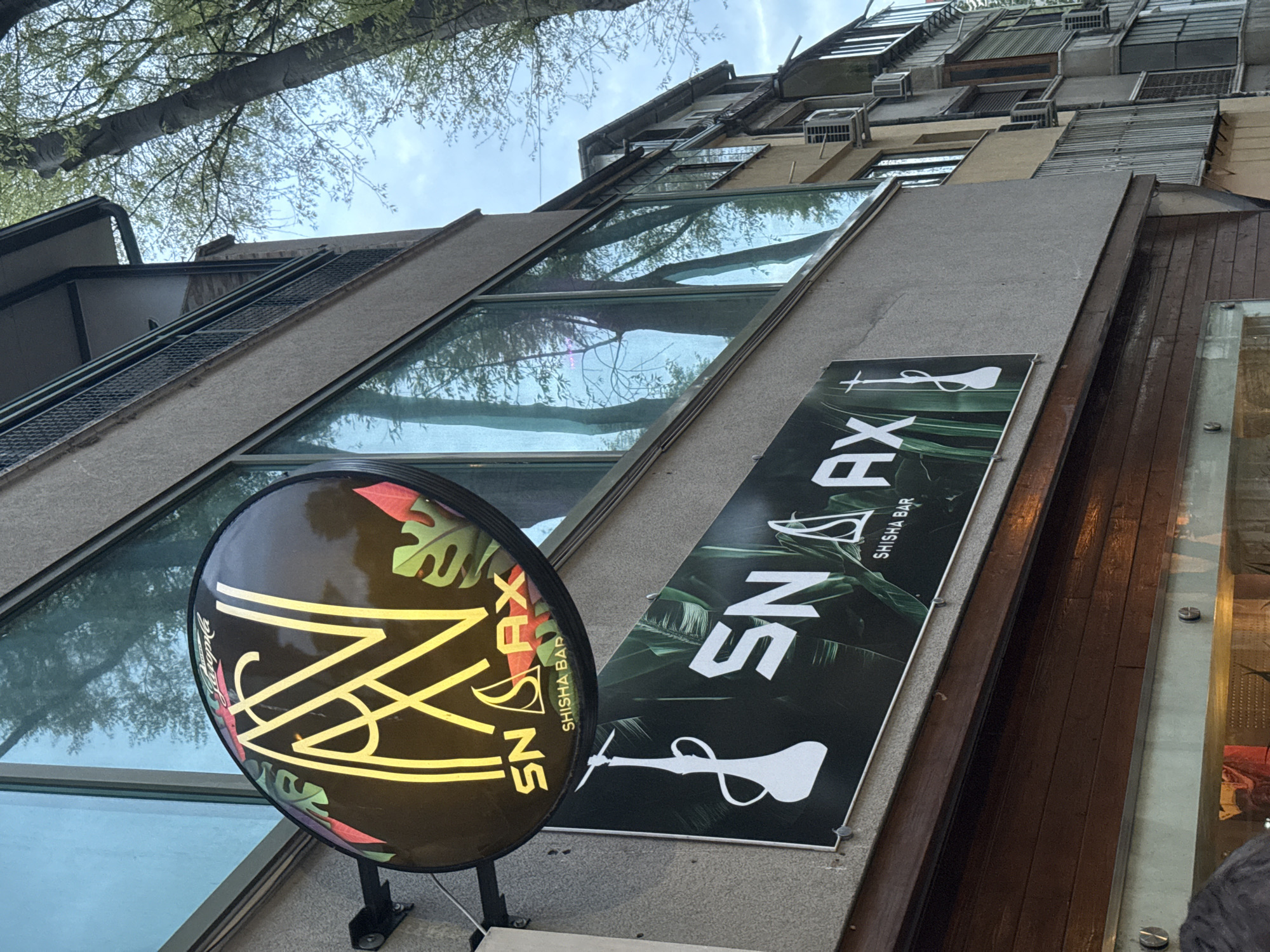 The image displays an upward-angled view of a building facade in Varna, Bulgaria, featuring two prominent signs for a "SN AX SHISHA BAR." The perspective captures the street-level and mid-sections of the structure, extending towards the sky and a tree canopy above.
A circular, illuminated sign prominently features the "SN AX" logo in yellow, abstract lines against a dark background with red and green tropical leaf motifs. Below the main logo, "SHISHA BAR" is also visible. Adjacent to it, a rectangular banner sign showcases "SN AX SHISHA BAR" in white text on a dark green background, complete with a dark tropical leaf pattern and a white silhouette of a shisha pipe.
The building incorporates large, angled glass windows that reflect the branches and light green foliage of a nearby tree, as well as the partly cloudy sky. The facade appears to be constructed from concrete-like materials, with sections of dark wooden paneling. Higher up on the building, numerous air conditioning units and various window fixtures are visible. The lighting suggests it is daytime, under an overcast or partly cloudy sky. No specific human activity is taking place; the image focuses on the architectural details and signage.