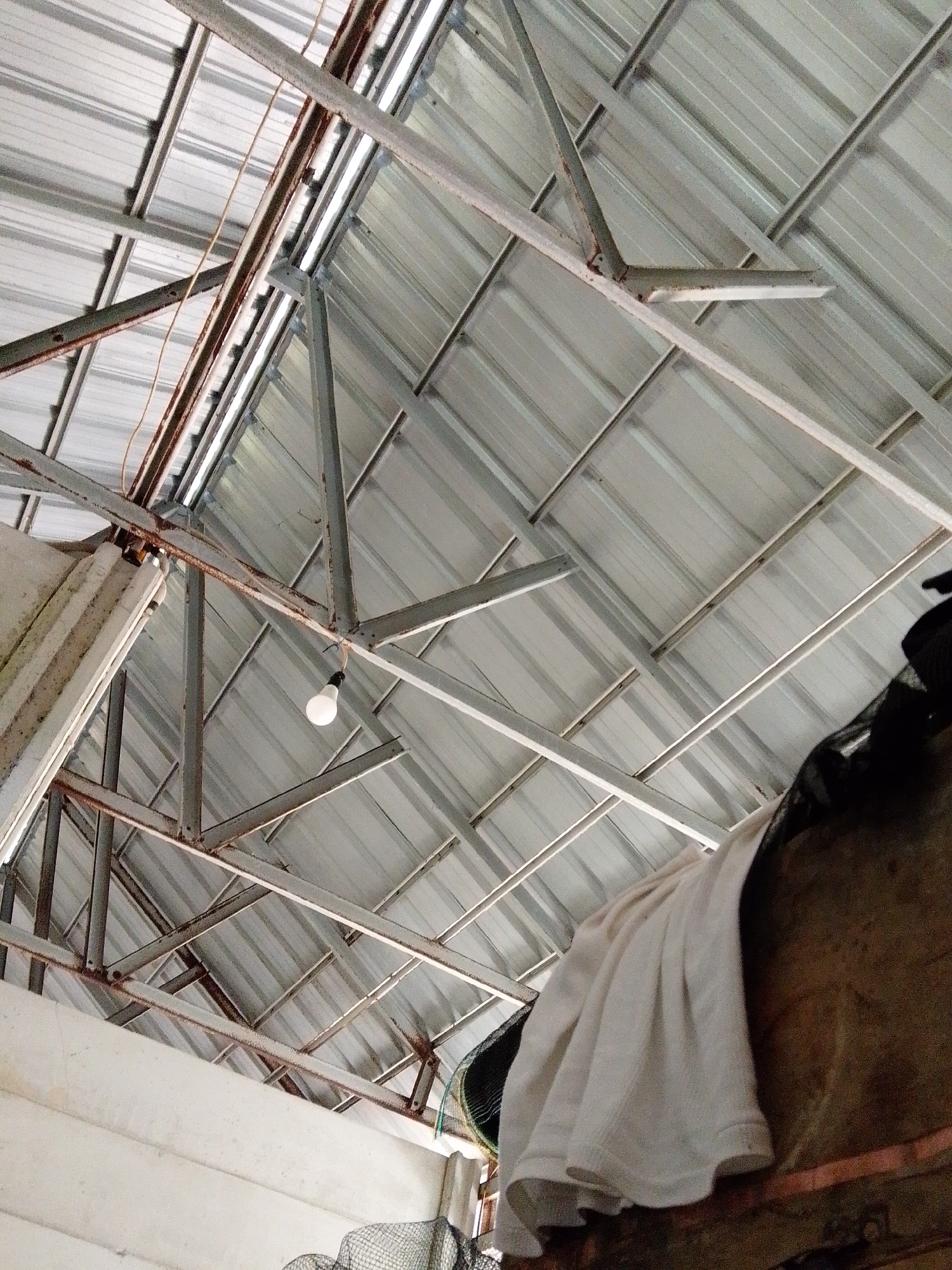 The image depicts the interior of a structure in Bukit Ketok, Indonesia, likely a shed or outbuilding, characterized by a metal roof with a network of support beams. A single lightbulb hangs from the structure. A white towel and a dark object are visible in the lower right corner of the image, obscuring part of the scene. A white wall with some items in front of it is seen on the left side of the image. The overall scene suggests a storage space or work area.