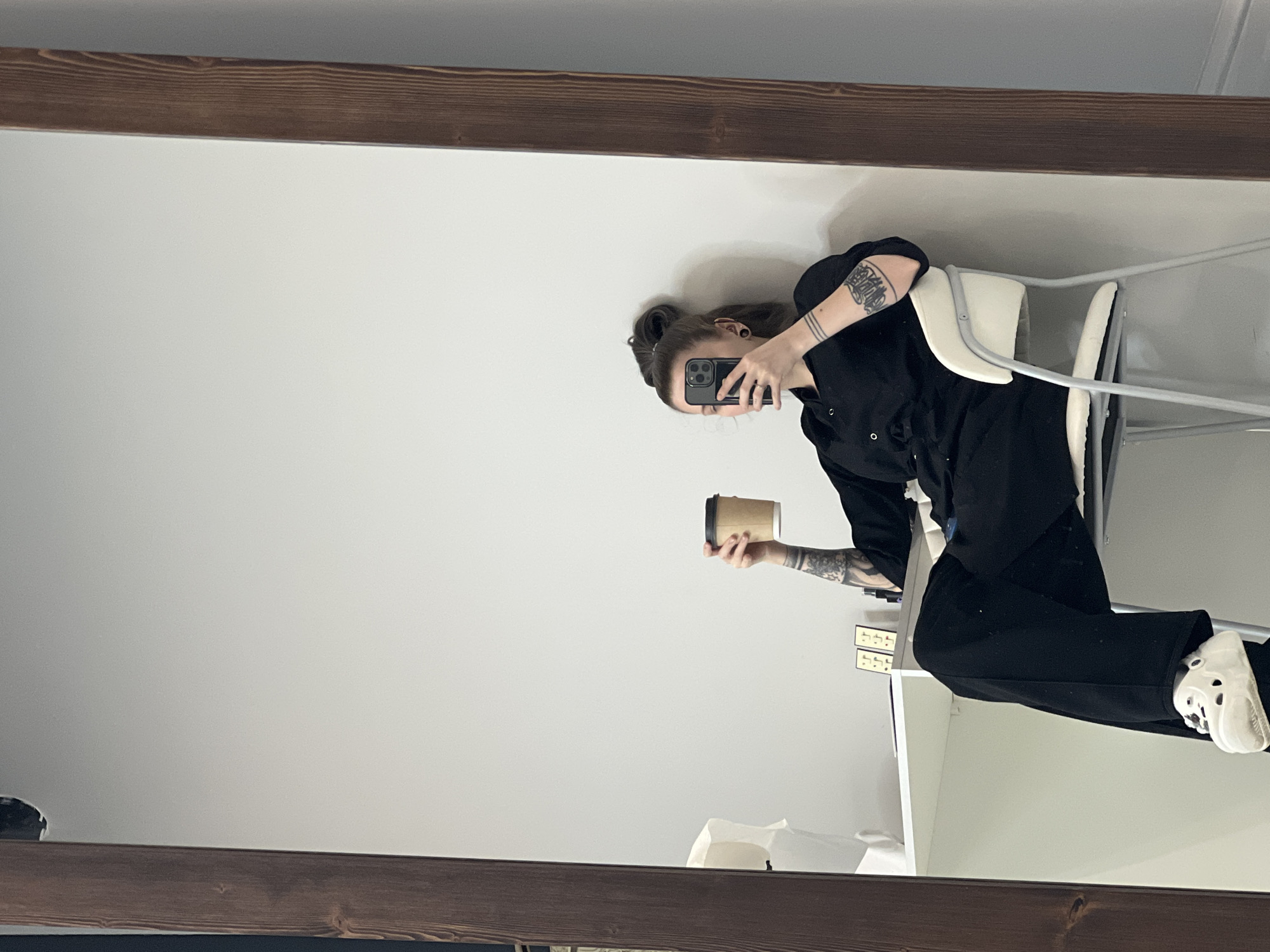 The image depicts the reflection of a female individual in a large, vertically oriented mirror with a dark brown wooden frame. The individual, seen from approximately the chest down to the feet, has dark hair styled in a bun, a black ear tunnel in her left ear, and multiple tattoos, including abstract designs on her left forearm and three horizontal lines on her right forearm. She is dressed in a black collared jumpsuit or two-piece outfit and white Crocs on her feet. She is seated on a white and light grey chair, likely a bar stool or high chair, with her legs crossed at the ankles.
Her right hand holds a black smartphone to her face, partially obscuring it, suggesting she is capturing her own image. Her left hand holds a kraft paper-colored disposable coffee cup with a black lid, positioned above her head. The background, visible in the reflection, consists of a plain white wall. To the reflected individual's left, a white shelf or desk is visible, holding two small, golden-colored rectangular decorative items and a white crumpled object. No specific visual details within the image allow for the identification of a precise location within the city of Khimki, Russia.