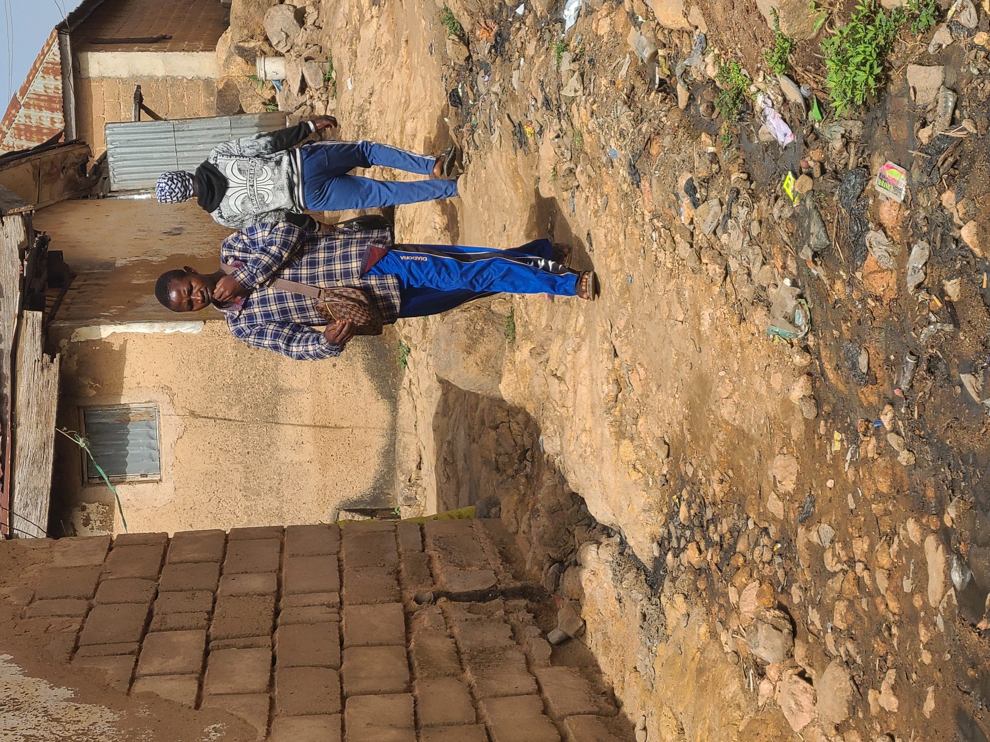The image depicts two individuals in Jos, Nigeria, standing on a rocky, dirty surface. The foreground figure wears a blue and black plaid shirt, blue sweatpants with "DIADORA" printed on the side, and sandals. He is holding a brown patterned bag and smiling. The background figure wears a gray and black sweatshirt, blue pants, and a white and blue cap. They are both facing the right side of the image.
The environment is littered with trash and rocks, and some green vegetation is visible. A building made of tan bricks and a corrugated metal roof is in the background on the left side. The overall impression is that of an impoverished area with poor sanitation.