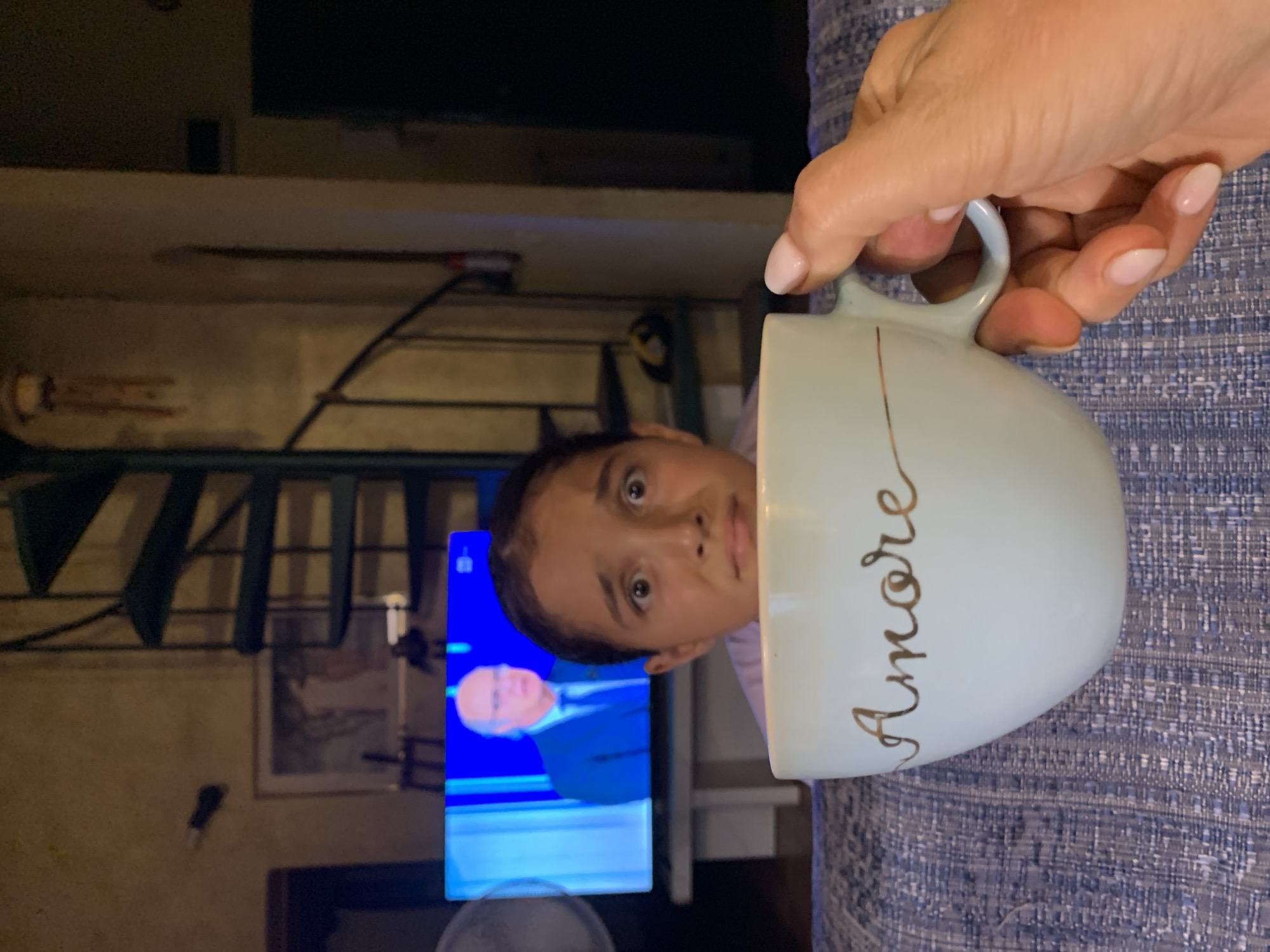In a dimly lit room, a young girl with wide, fearful eyes peeks over a light blue coffee mug that has "Amore" written in gold script. She is seated on a patterned sofa, with a television screen in the background displaying a man in a suit and tie. A spiral staircase is visible behind the television. A hand with manicured nails is holding the mug. The overall ambiance suggests a domestic setting, possibly in a town like San Quirico d'Orcia, Italy.