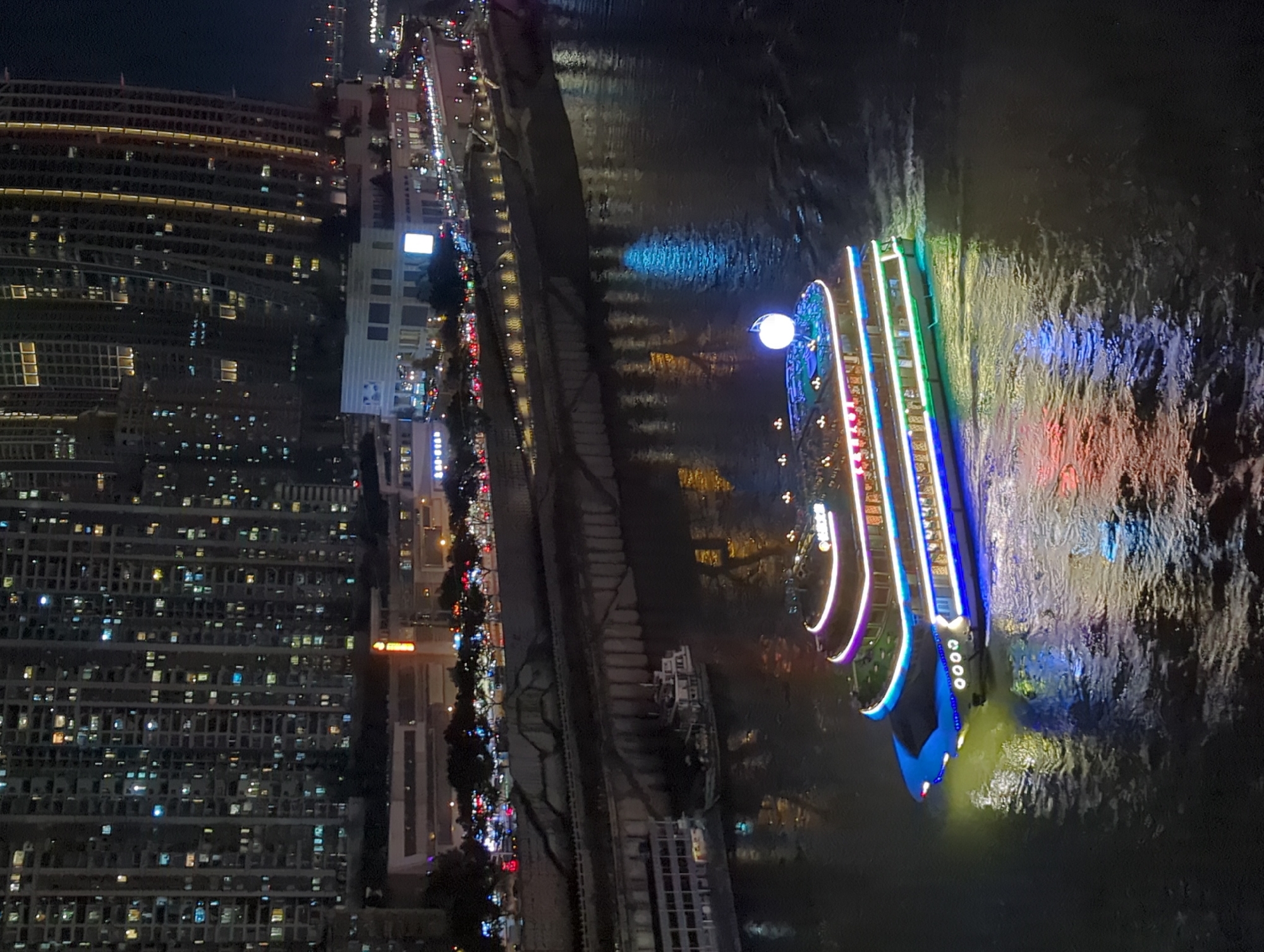 An elevated nighttime aerial view captures the Yuzhong District of China, a bustling urban landscape dominated by towering, illuminated skyscrapers. The buildings, densely packed, feature numerous windows, many of which glow with warm interior lights, creating a textured facade against the dark sky. Below the skyscrapers, a multi-level roadway is visible, marked by the red taillights of numerous vehicles, indicating heavy traffic flow. Along the riverbank, a waterfront promenade is illuminated with decorative lights. A large, illuminated tour boat is the primary subject in the foreground, navigating the dark, reflective waters of a wide river. The boat is adorned with vibrant LED lights in shades of blue, green, purple, and yellow, which cast colorful reflections onto the water's surface. Its structure features multiple decks with visible windows and railings. Another, smaller boat is docked near the riverbank in the middle ground. The overall scene depicts a lively and visually dynamic urban environment at night.