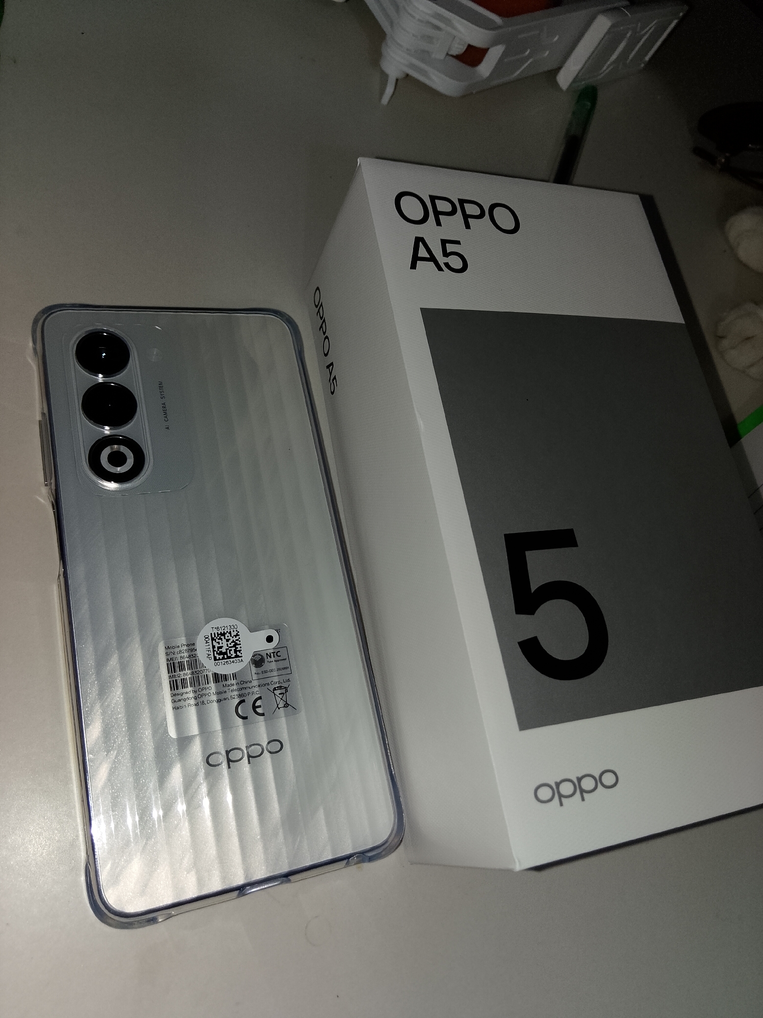 The image displays a new OPPO A5 smartphone placed next to its retail packaging. The phone, silver in color with a textured back, is partially covered by a clear plastic protective film. Its rear camera module, featuring three circular lenses, is visible. A sticker on the back of the phone shows various labels including "Mobile Phone," "S/N," "IMEI," and a QR code, alongside regulatory marks like "CE" and "NTC Type Approved." The OPPO A5 packaging is white with black text, prominently featuring the brand name and model number, as well as a large black number "5" on a grey panel. The overall setting appears to be an indoor environment, likely a desk or table, with a neutral-colored surface. The lighting is somewhat dim, creating shadows and highlighting the reflective surfaces of the phone and packaging. The presence of the NTC sticker indicates the device is intended for the Philippine market. Given the NTC approval, this product is likely being sold or used within the Philippines, and potentially within Davao City.