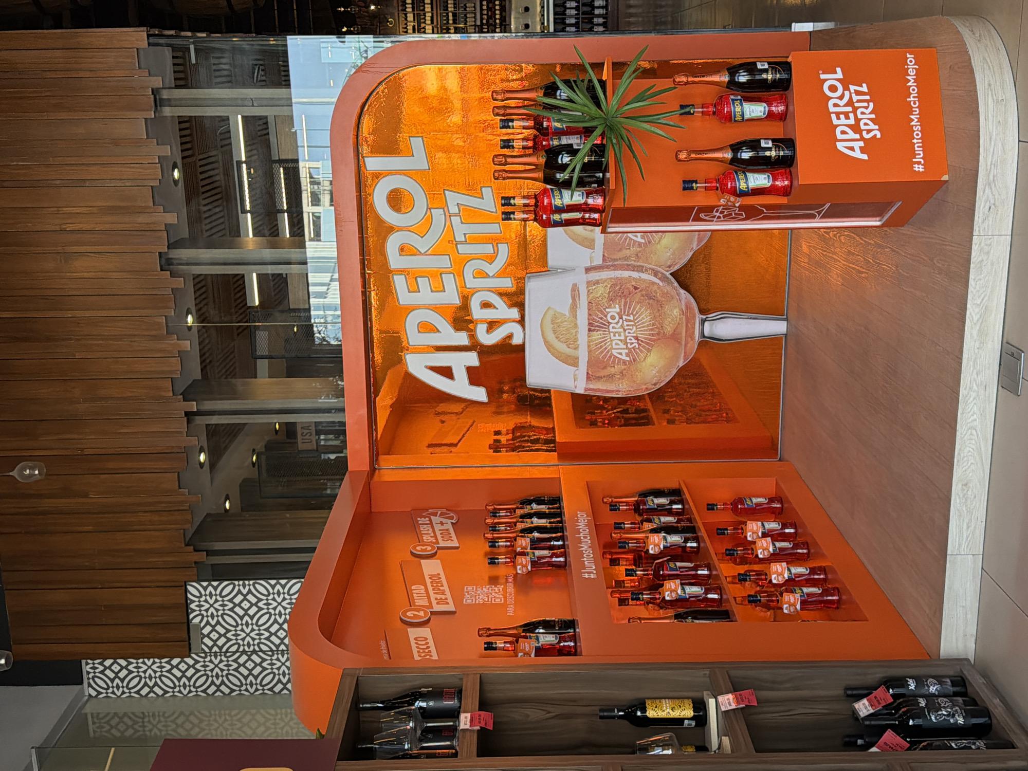 This image captures an indoor retail display for Aperol Spritz, oriented sideways, likely in a liquor store or supermarket in Puebla, Mexico. The central subject is a vibrant orange Aperol Spritz promotional stand, prominently featuring large white "APEROL SPRITZ" lettering and a graphic of a cocktail glass filled with the drink and an orange slice.
The display is well-stocked with numerous small bottles of Aperol and what appear to be bottles of sparkling wine (likely Prosecco), arranged on multiple tiers. An artificial green plant adds a decorative touch on the top shelf. One section of the display clearly outlines the recipe for an Aperol Spritz with numbered steps: "1 SECCO" (Prosecco), "2 MITAD DE APEROL" (half Aperol), and "3 SPLASH DE SODA" (splash of soda). Below these instructions, a QR code is visible with the text "PARA DESCUBRIR MÁS" (to discover more). The hashtag "#JuntosMuchoMejor" (Together Much Better) is featured prominently on different parts of the display.
In the foreground and background, other store shelves are visible, displaying various bottles of wine and spirits. Reflections in a glass surface show architectural details like wooden paneling and lighting fixtures. Partial text like "USA" and "ana de hiel" can be seen in reflections and on other products. The overall setting is brightly lit, suggesting daytime inside a commercial establishment.