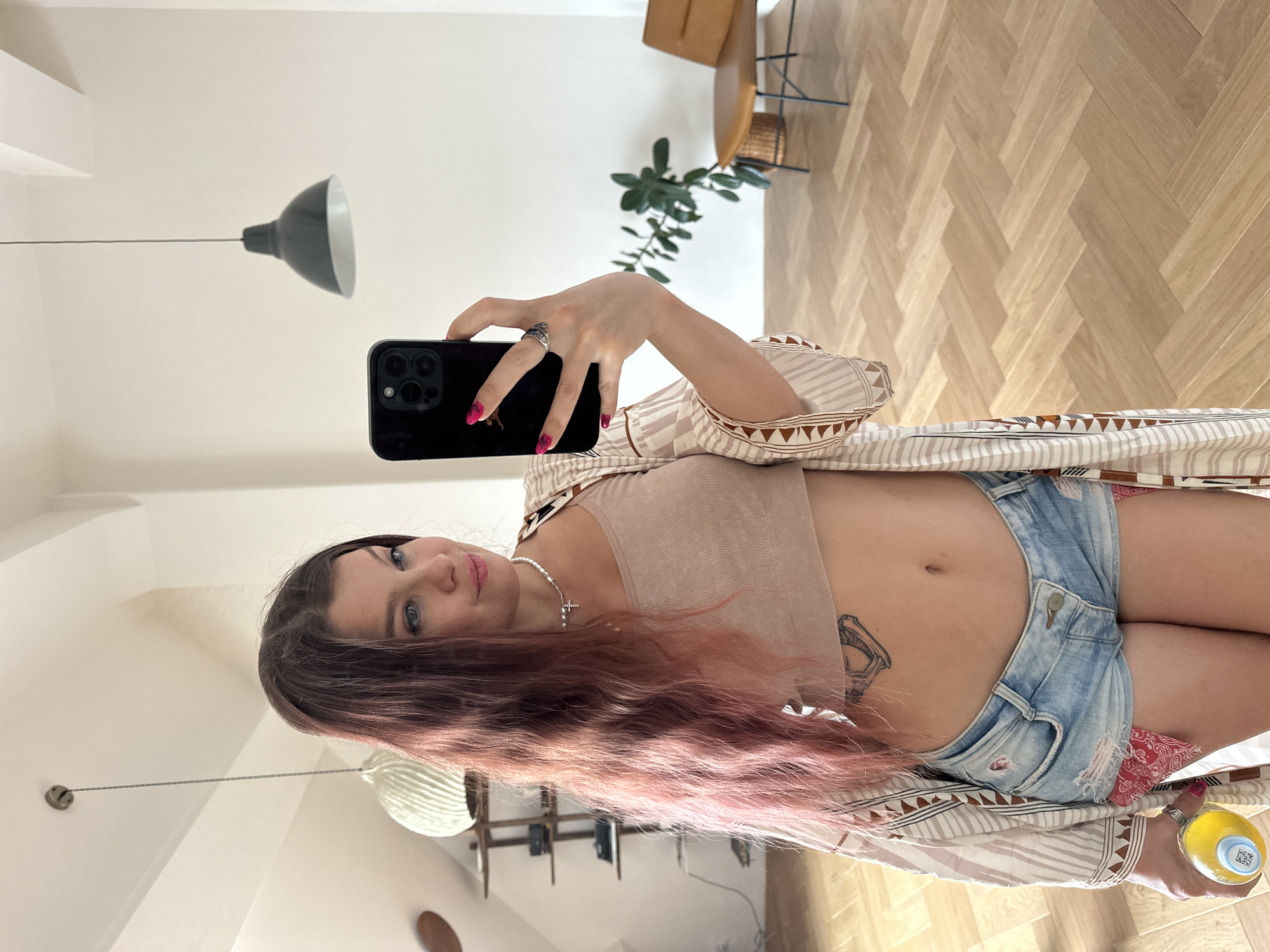A young woman with long, wavy, pink-tinted hair takes a mirror selfie in a well-lit room. She is wearing a light beige crop top, distressed denim shorts with a red bandana peeking out from the pockets, and a patterned open-front kimono. A silver necklace with a cross pendant rests on her collarbone, and a silver ring is on her right index finger. She is holding a yellow bottle in her left hand and a black smartphone in her right hand, capturing her reflection. The room features a light-colored wooden floor laid in a herringbone pattern. In the background, a brown leather chair sits near a potted plant, and shelves with various items are visible on the wall. Two ceiling lamps hang in the room. The scene appears to be an indoor residential space.