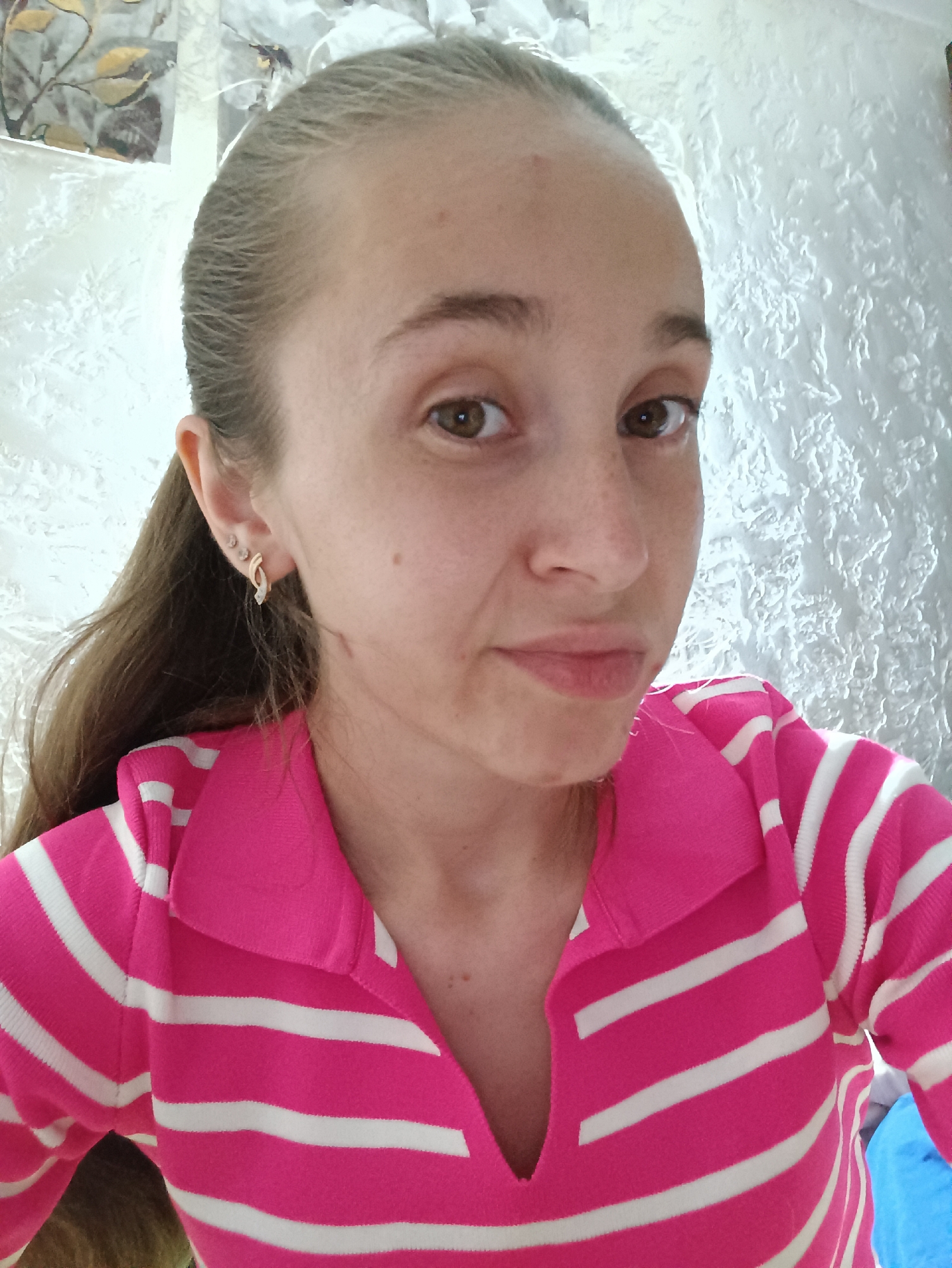 A young Caucasian woman with blonde hair pulled back in a ponytail is captured in a close-up, head-and-shoulders portrait. She is wearing a pink and white striped shirt with a collar and a V-neck. Her expression is neutral, with her eyes looking directly at the camera. She has subtle makeup and is wearing earrings. The background appears to be an indoor setting with textured walls and a patterned element to the left. The lighting is soft, illuminating her face and upper body. The image is a selfie taken by the subject. The location information provided is specific to Vypasne, Ukraine, however, no visual cues within the image itself corroborate this location.