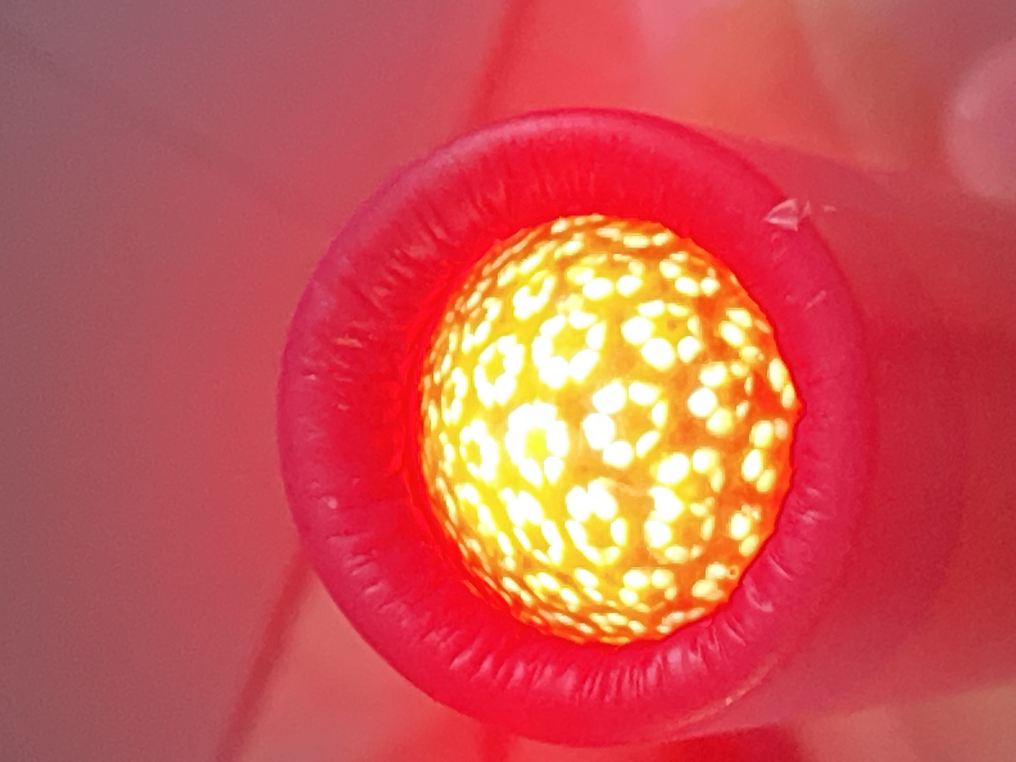 The image shows a close-up view of a bright yellow spherical object with a patterned surface, appearing to be illuminated from within. This object is enclosed by a vibrant red cylindrical structure. The background is a blurry gradient of pink and red. There is no discernible location in the city of Belyye Berega, Russia depicted in this image. The image does not contain any people, actions, or interactions.