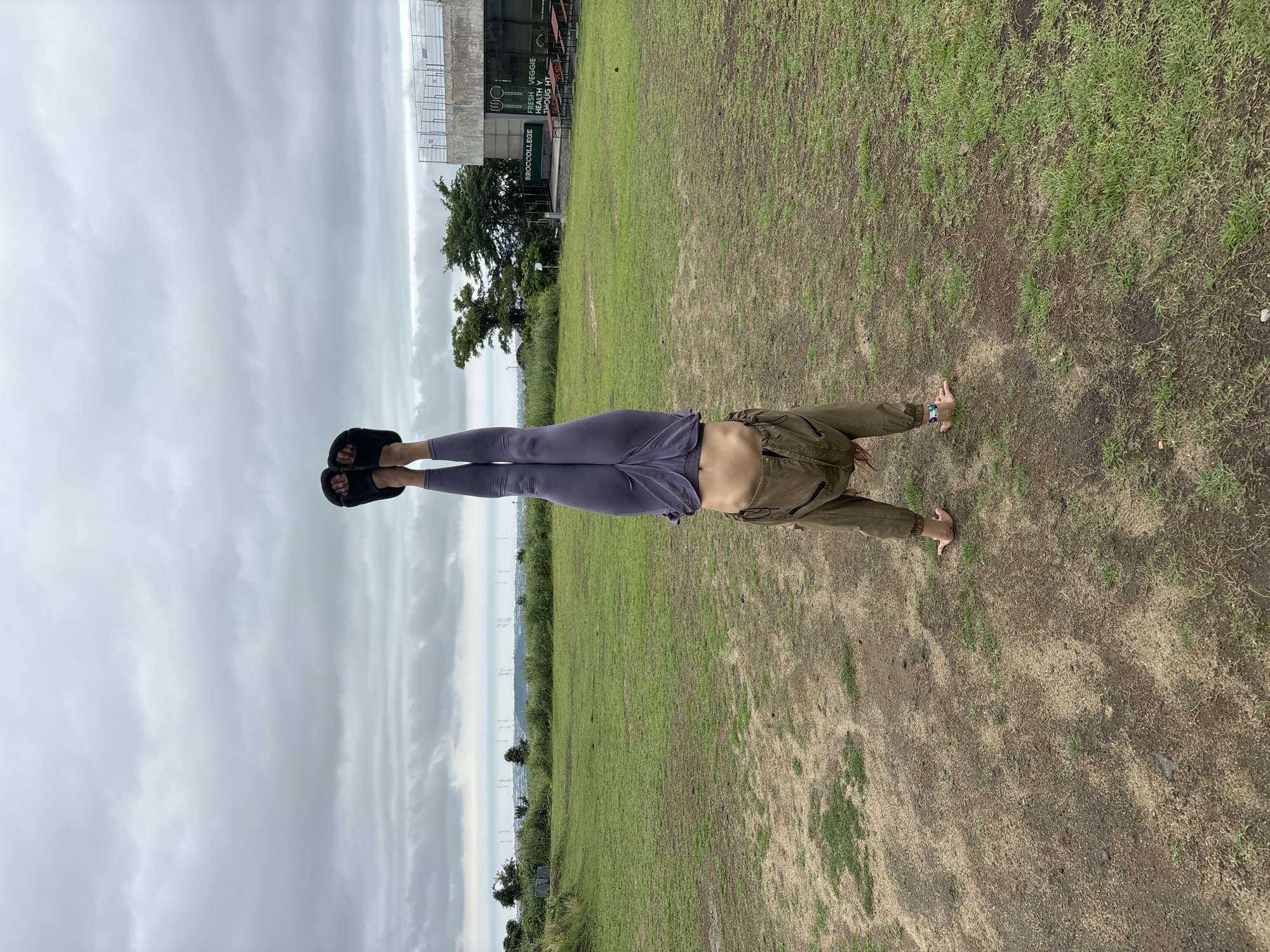 A person is performing a handstand with their legs extended straight in the air, creating a horizontal line. They are on their hands, with their body perpendicular to the ground. Their legs are clad in purple leggings, and they are wearing black slides on their feet. The person is wearing a brown jacket over a visible midriff. The setting is an outdoor grassy area, likely a park or field, with patches of dry soil. In the background, there is a modern building with signage, a lone tree, and a cloudy sky. The overall weather appears overcast. The image's orientation is landscape, with the person positioned vertically within the frame. The camera angle is from a low perspective, looking up towards the person. No specific time of day is discernible, but the cloudy conditions suggest it could be morning or afternoon. The location is identified as Jeju, South Korea. The visible text on the building reads "BROCCOLLEGE" and "FRESH VEGGIE HEALTHY THOUGHT." The person's pose suggests athleticism and concentration.