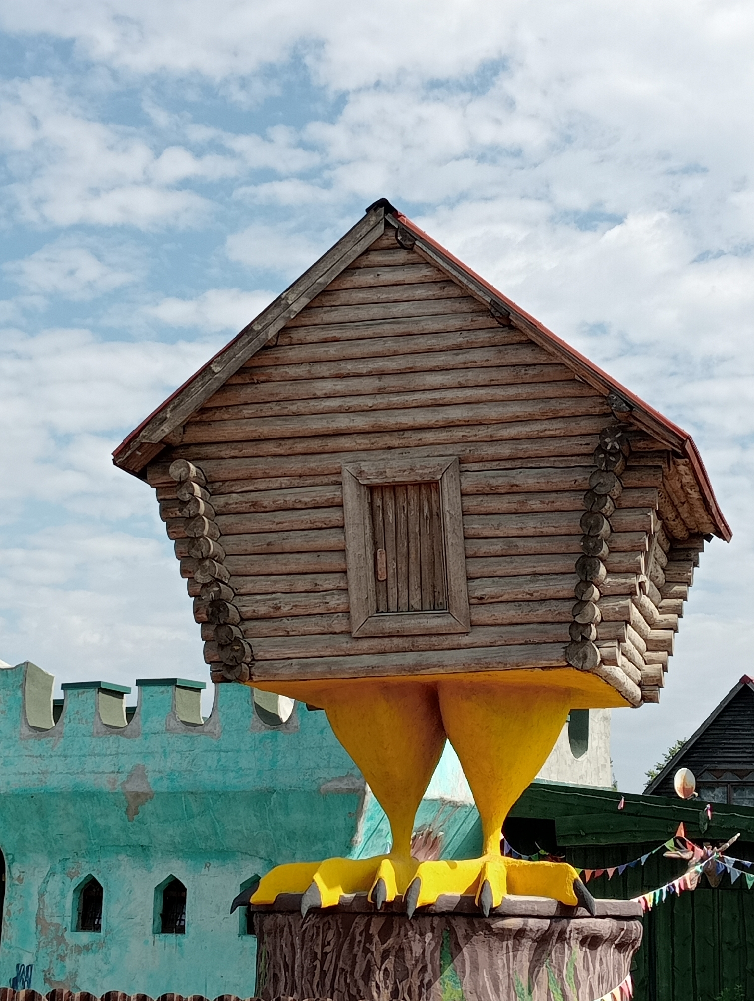 A whimsical wooden structure resembling a house with large yellow bird-like legs stands on a carved base. The house has a red roof and a single wooden door on its facade. In the background, a turquoise wall with crenellations and arched windows suggests a park or amusement setting. A wooden fence with colorful bunting is visible in the foreground and to the right. The sky is partially cloudy. The location is specified as the city of Ulyanovka, Russia, likely within a park or entertainment venue, given the playful architecture. No people are visible in the image.