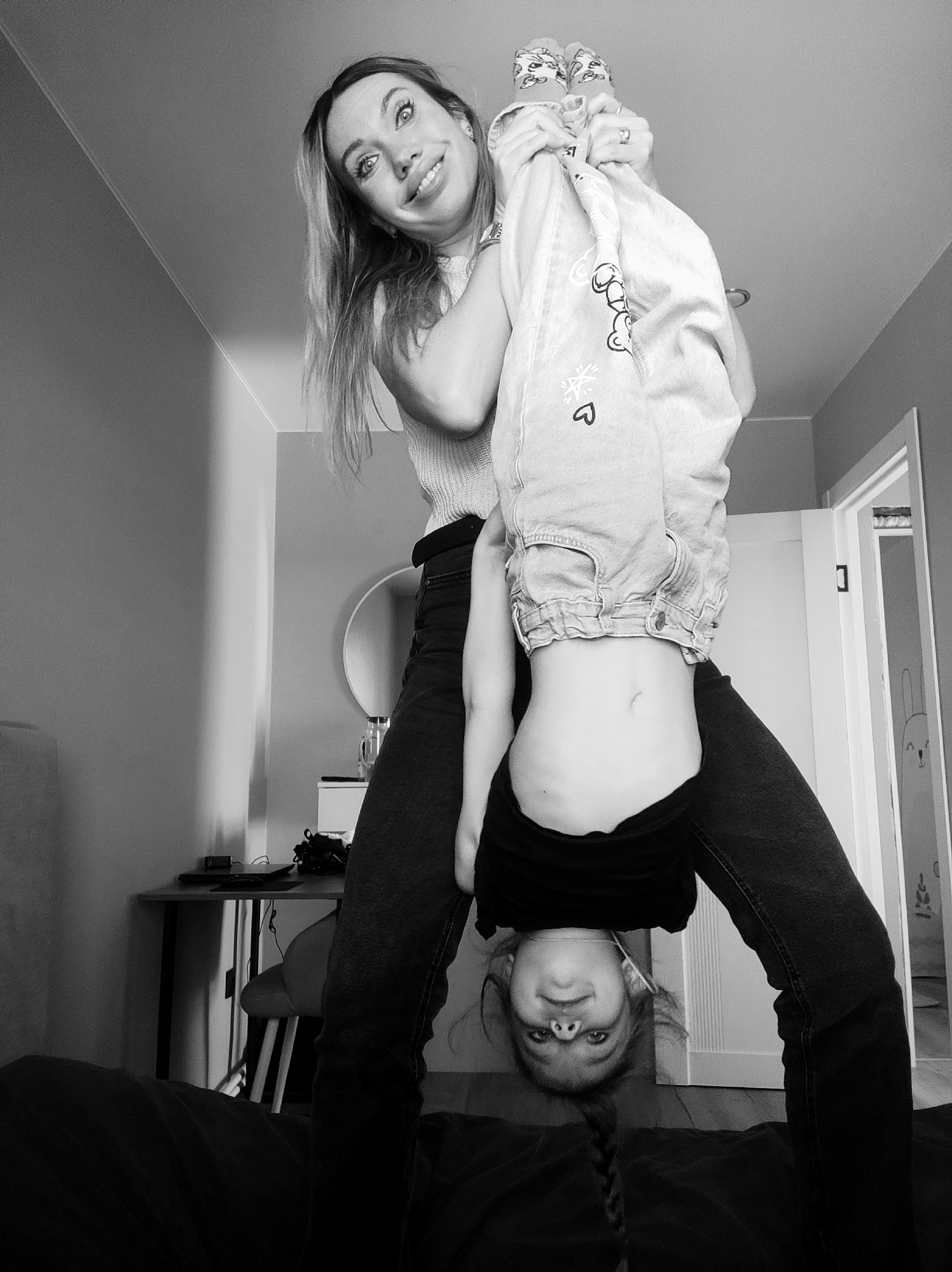 A black and white, low-angle shot depicts a woman holding a child upside down in what appears to be an indoor setting. The woman is smiling and looking towards the camera while holding the child by the ankles. The child is clad in light-colored jeans with cartoon drawings, a dark top, and is also looking towards the camera with a smile.
The location appears to be a domestic space, possibly a bedroom or living area, with a bed in the foreground and a desk with electronic devices in the background. A round mirror is visible on the wall. A door is ajar revealing a drawing on the wall inside the next room, possibly a child's drawing.
The exact location within the city of Votkinsk, Russia cannot be determined from the image alone.