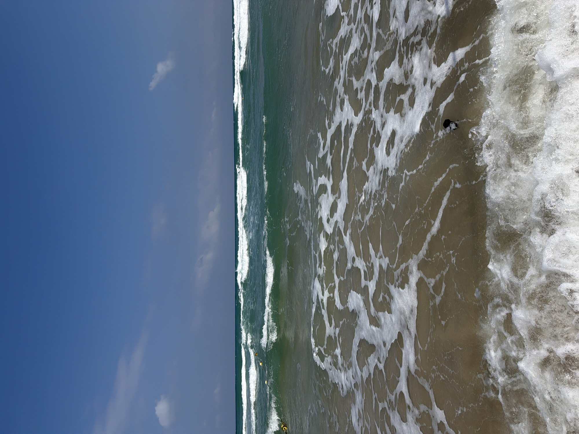 The image depicts a serene beach scene in Ashdod, Israel. The foreground is dominated by the gentle lapping of waves against the sandy shore, creating intricate patterns of white foam on the translucent water. In the middle ground, the sea displays a striking emerald hue, with more pronounced waves rolling in, their crests capped with white foam. Further out, the horizon is marked by a line of breaking waves, indicating a moderate surf. The sky above is a vast expanse of clear blue, punctuated by a few wispy white clouds. In the distance, on the water, several yellow buoys are visible, potentially marking a swimming area or a navigational point. A small black object, possibly a piece of debris or a stray item, floats in the water near the shore. The overall impression is one of tranquility and natural beauty.
