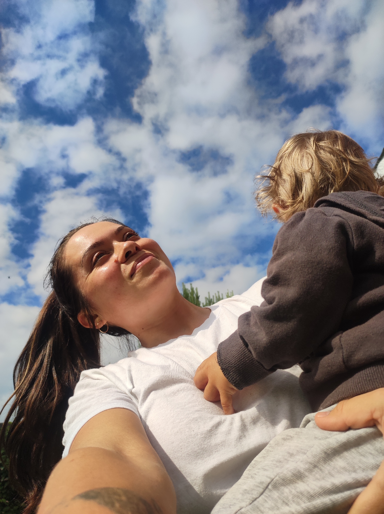 In an outdoor setting under a cloudy blue sky, a young woman with long, dark hair pulled into a ponytail is holding a baby. The woman is wearing a white t-shirt and a small gold hoop earring. She is looking up and smiling, with sunlight illuminating her face. The baby, whose back is to the camera, has blond, curly hair and is wearing a dark hooded sweatshirt and light grey pants. The baby's hand is resting on the woman's chest. The image is taken from a low angle, looking up towards the sky. The environment appears to be a natural setting with some greenery visible at the bottom of the frame. No specific landmarks or elements indicating the city of Фоки, Russia, are present in the image.