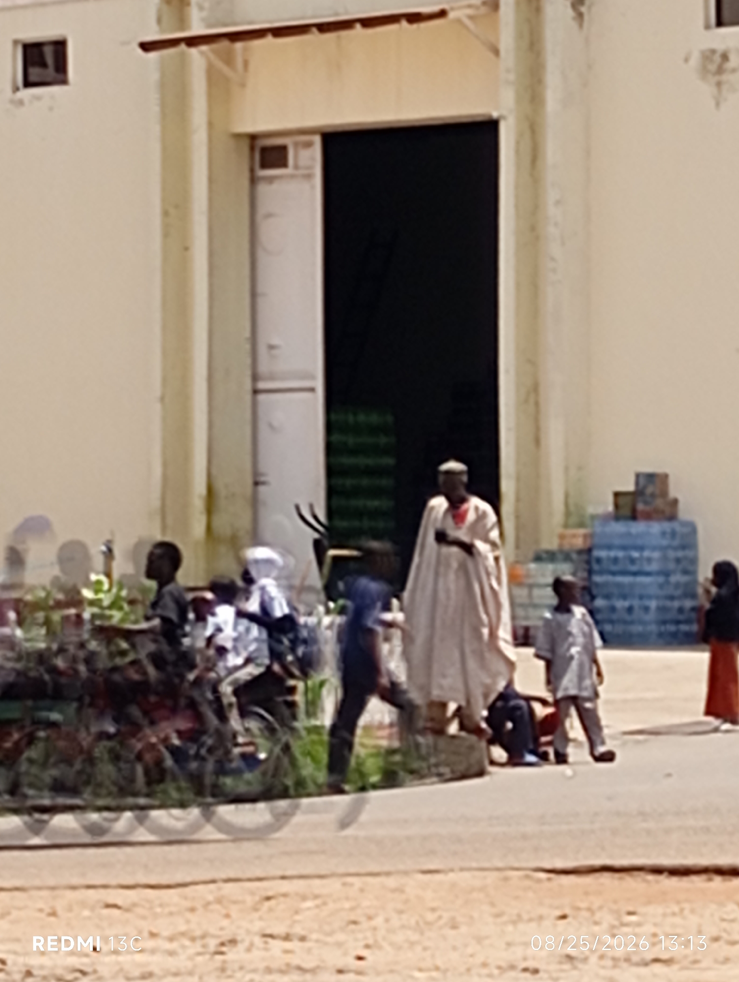 The image shows a group of people outside a building with a large open doorway. In the foreground, a blurred motorcycle with riders is visible. To the right, stacks of blue bottled water are arranged. A man in a white robe and head covering stands in the middle ground, facing left. Several children are also present, some appearing to be interacting with the man. The environment appears to be a dusty, outdoor area next to a commercial building, possibly a warehouse or distribution center. The timestamp "08/25/2026 13:13" is present in the lower right corner.