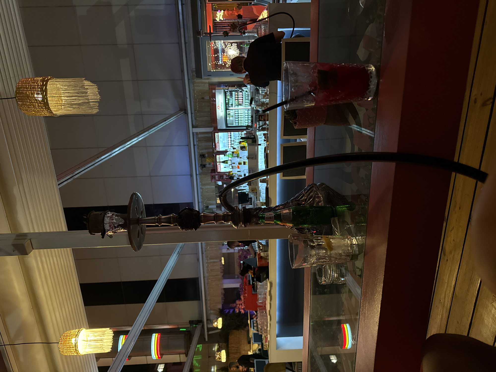 This media file captures a vibrant night scene at an outdoor or semi-outdoor lounge area in Plovdiv, Bulgaria, viewed from a patron's perspective. The general setting suggests a bar or restaurant operating in the evening.
In the foreground, a dark shisha (hookah) with a long, curved black hose sits on a glass-topped table. Adjacent to it, a clear glass filled with a red drink and ice, complete with a dark straw, is positioned on a dark reddish-brown counter or table extension. An empty glass and a green bottle are also visible near the shisha base.
Two distinctive cylindrical pendant lamps, with woven golden exteriors and fringed bottoms, illuminate parts of the scene, hanging from the ceiling structure.
The midground reveals a bustling bar area. A person, seen from behind, is actively working at the bar counter. Behind them, shelves are densely packed with various bottles of alcohol. Two large white refrigerators are visible, stocked with beverages, and one prominently features "Plovdiv" written on its front, confirming the given location. The bar counter is brightly lit from beneath with a cool, possibly blue or white, light strip.
In the background, other patrons are dimly discernible, enjoying the atmosphere. Additional decorative lighting includes a small, colorful, striped neon-like light showing red, yellow, and blue. The ceiling structure appears to be a white and grey paneled canopy or tent, indicating a covered outdoor space. The overall ambiance is casual and social, typical of an evening gathering spot.