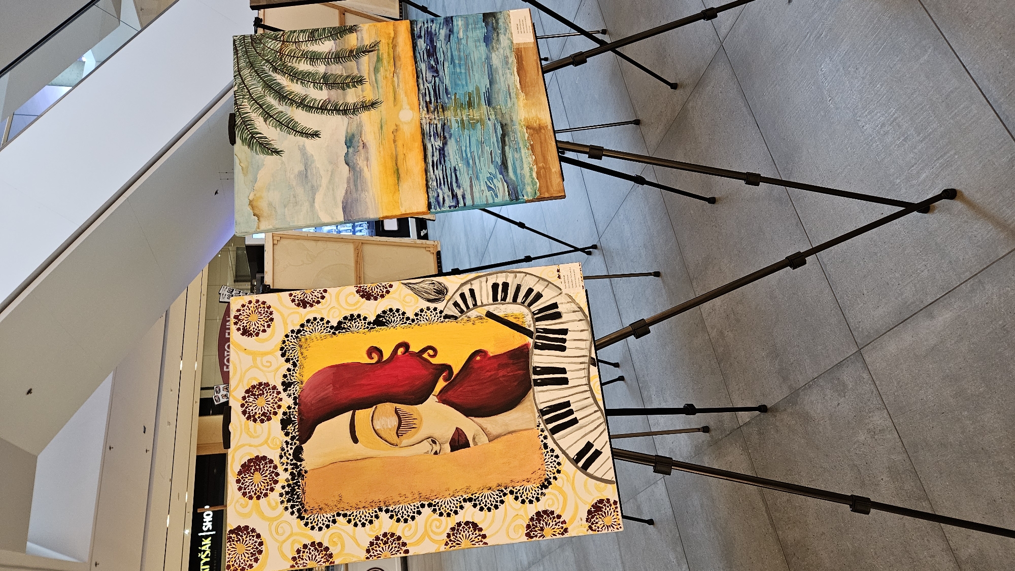 An indoor art exhibition is in progress, featuring two prominently displayed paintings on easels. The painting on the left, rendered in yellow, depicts a woman's face with closed eyes, partially obscured by her deep red hair, and is complemented by a black and white piano keyboard design. The painting on the right is a landscape of a tropical beach at sunset, with a palm tree in the foreground, a calm sea reflecting the warm sky, and mountains in the distance. The exhibition is located in the shopping mall OC Eperia in Prešov, Slovakia, as indicated by the visible signage for "ATYŠÁK | SHOP" and "FOTO FUN". The floor is tiled with large, light grey square tiles. The lighting suggests a well-lit indoor environment, possibly natural light from a skylight or large windows.