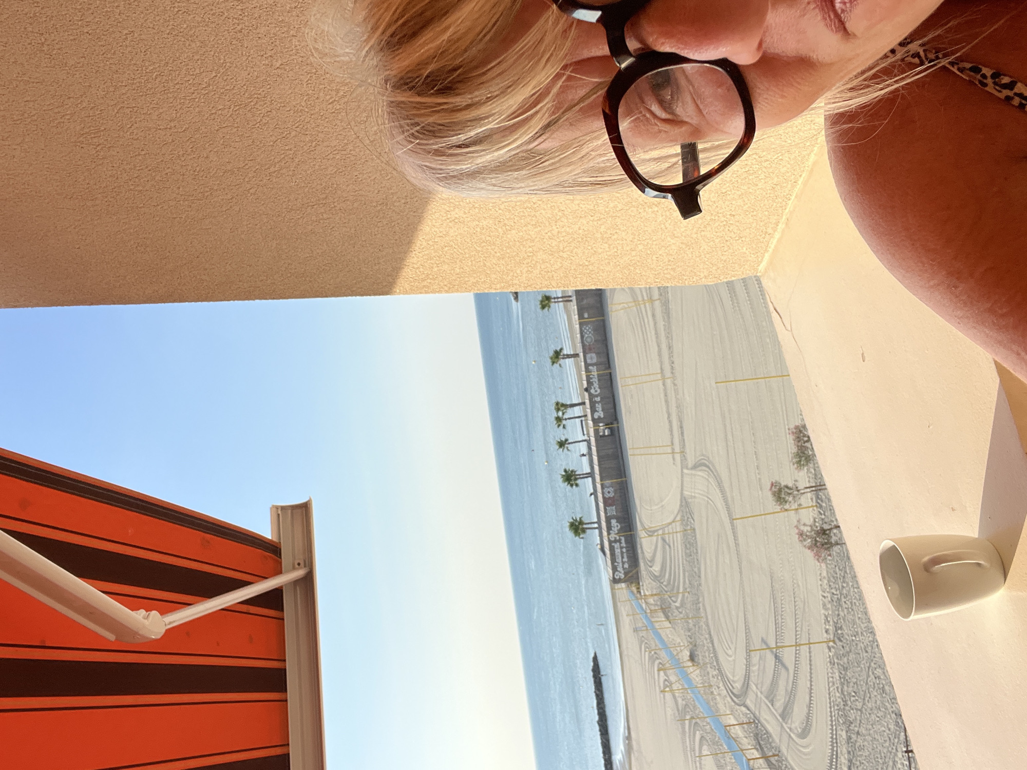 A woman with blonde hair and glasses is looking at the camera from the right side of the image. She is wearing a leopard print top. She is on a balcony overlooking a beach in Palavas-les-Flots, France. A white mug is on the railing in front of her. The beach has rows of sun loungers, palm trees, and a beach restaurant. The sea is blue and the sky is clear. An orange and brown striped awning is visible at the top of the image.