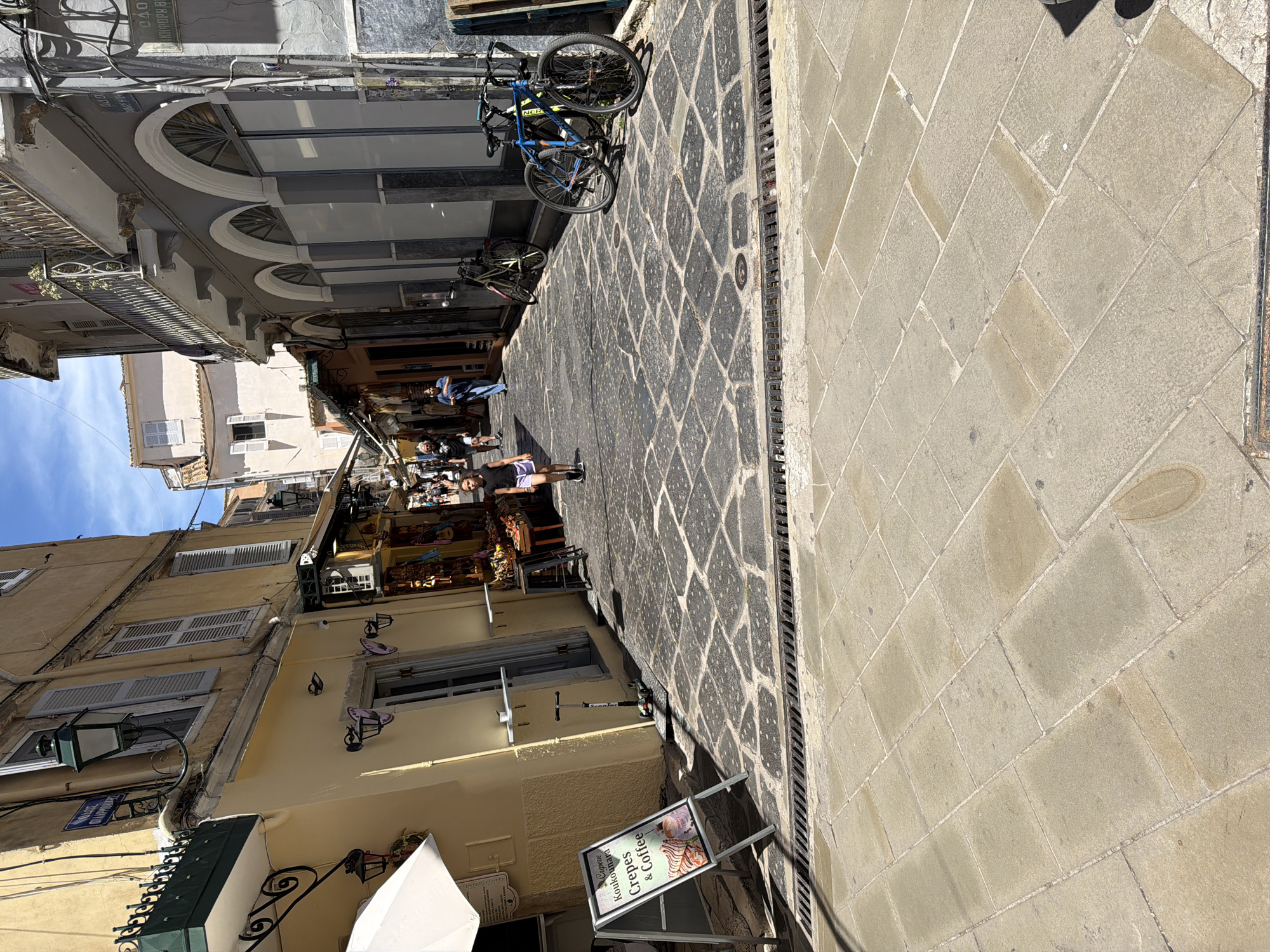 This image captures a vibrant, sunlit street or alleyway in Corfu, Greece, characterized by traditional architecture and a pedestrian-friendly atmosphere. The street is paved with a distinctive dark, patterned stone pathway, flanked on one side by a lighter-colored sidewalk made of rectangular stone slabs, separated by a long metal drain.
Buildings line both sides of the narrow passage. On the left, facades of warm yellow and white feature windows, some with closed white shutters, and wrought-iron balconies. A green street lamp and a blue street sign, partially visible, display "ΟΔΟΣ ΚΑΛΑΜΟΥ" (Kalamou Street) and the number "40" or "400." On the right, buildings exhibit a gray facade with multiple arched windows adorned with decorative grilles. Several bicycles, including a prominent blue one, are parked against these buildings.
The scene depicts everyday activity during a clear, sunny daytime. People are present on the street; one individual in a black t-shirt and purple shorts stands in the mid-ground, while others are visible further down the alley, suggesting a mix of residents and tourists. A shop displaying various goods adds to the commercial character of the street.
In the foreground, an A-frame sign prominently advertises "Crepes & Coffee" with the name "Koukounara Creperie." A white umbrella is also visible, likely providing shade for a cafe or shop. The overall impression is that of a charming, bustling European old town street.