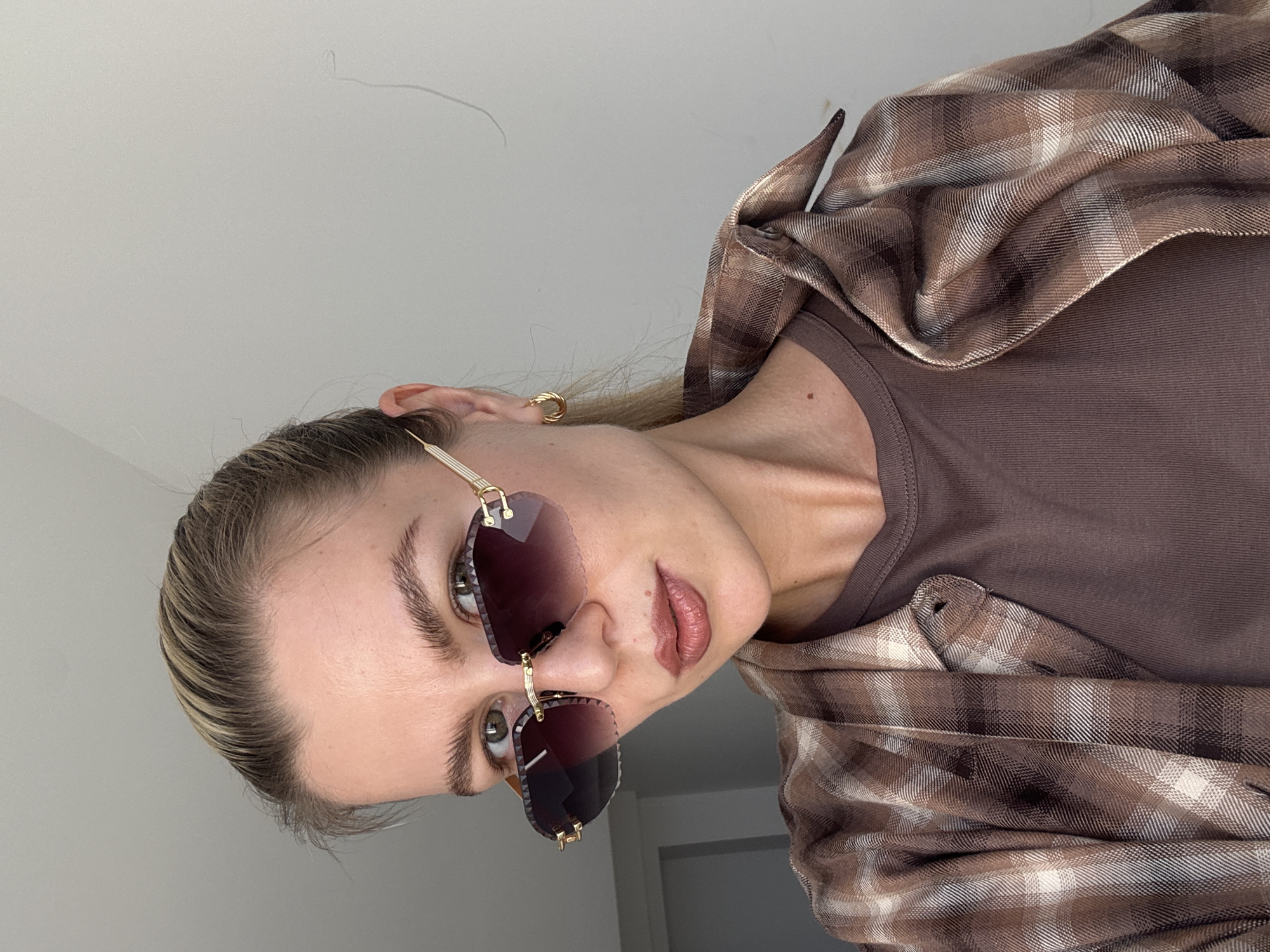 A woman is taking a selfie, looking at the camera. She has blonde hair pulled back into a ponytail and is wearing oversized sunglasses with a gold frame and tinted lenses. She is also wearing a brown plaid shirt over a brown t-shirt, and small gold hoop earrings. The background is a plain, light-colored wall.