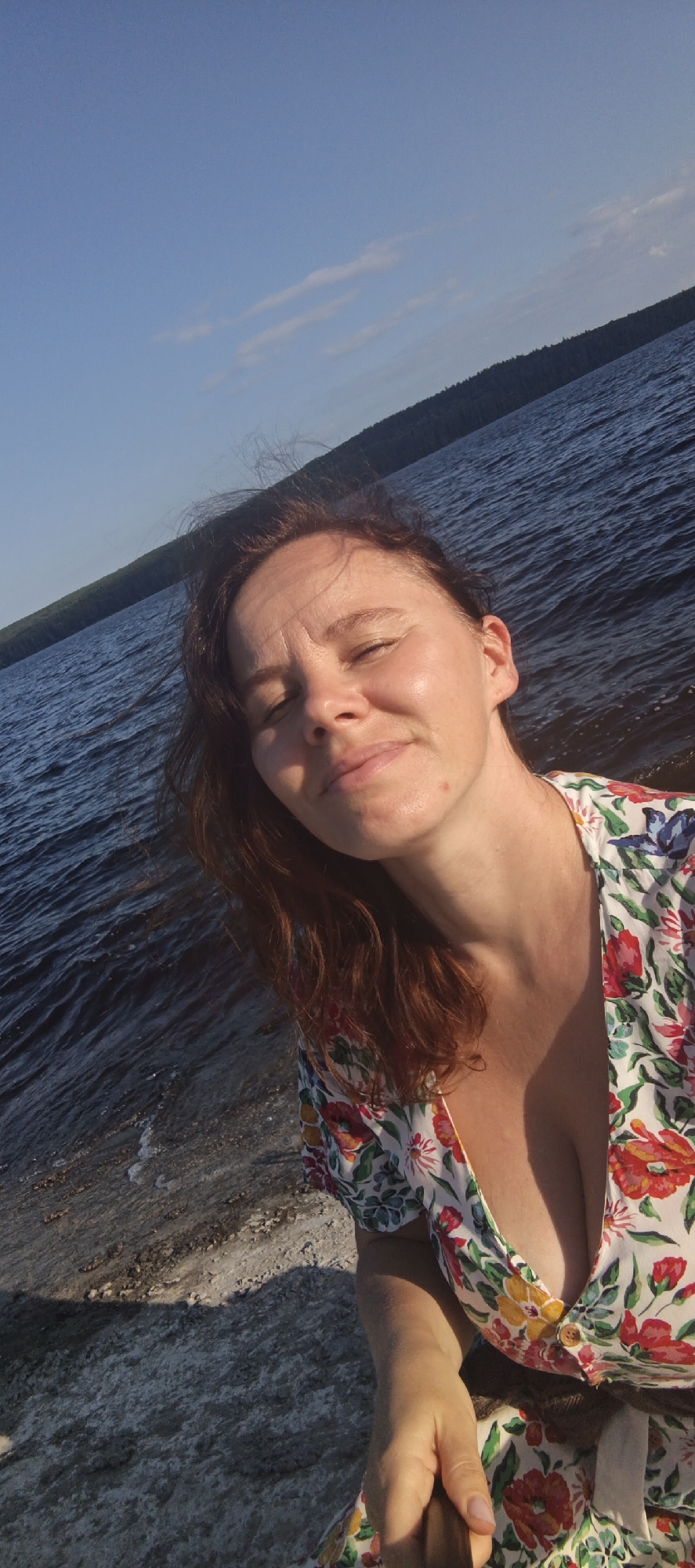 In this selfie, a woman with auburn hair and fair skin gazes at the camera with closed eyes and a slight smile. She is wearing a white dress adorned with a vibrant floral pattern featuring red, yellow, and pink flowers. The image captures her from the chest up, with the background showcasing a body of dark water under a clear blue sky. The sun appears to be low in the sky, casting a warm glow on her face, suggesting it is either early morning or late afternoon. A distant, tree-lined shore is visible on the horizon. The water is rippling, indicating a gentle breeze. The overall impression is one of tranquility and enjoyment of nature.