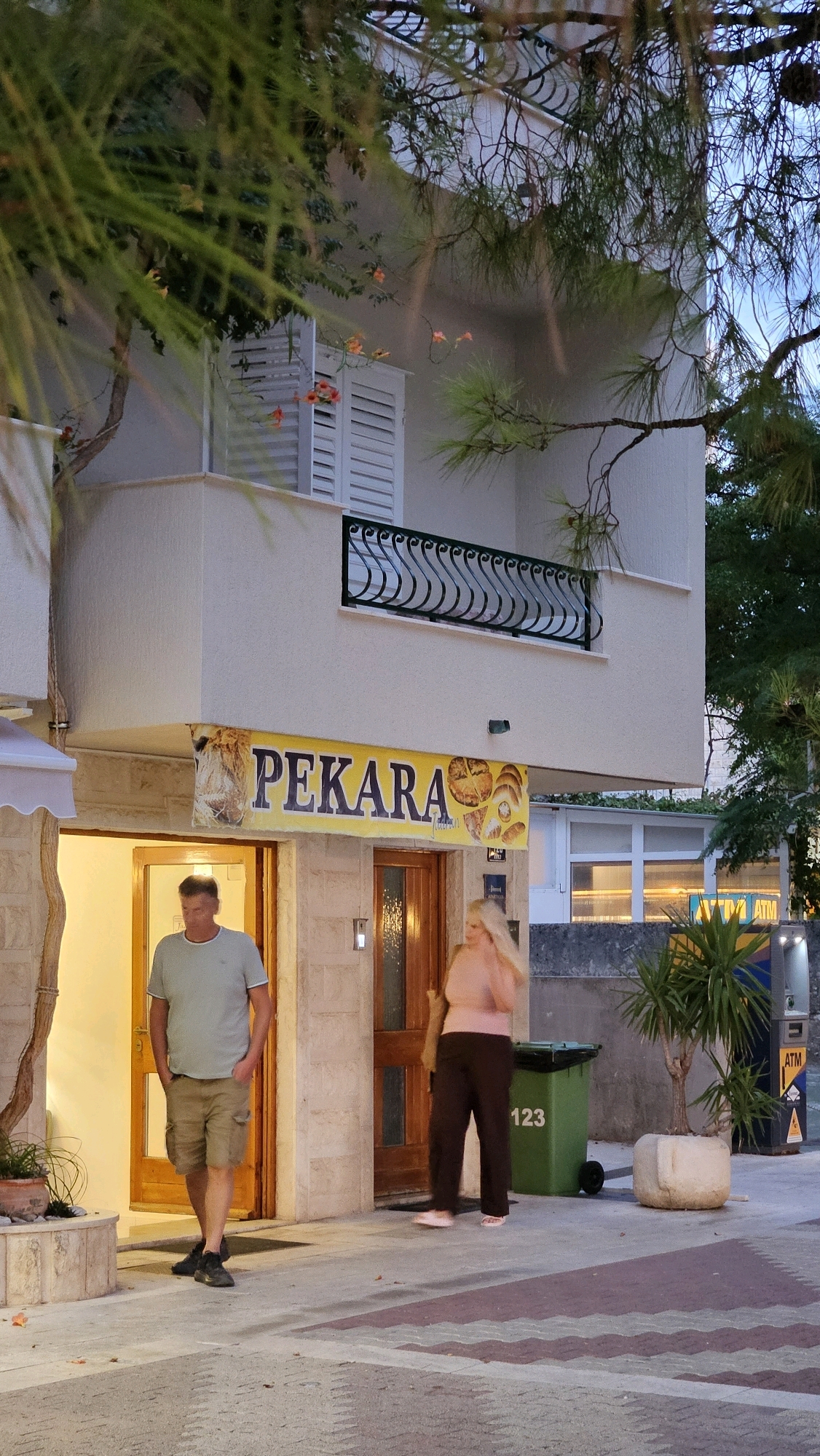 In Općina Tučepi, Croatia, a street-level view captures a bakery named "PEKARA" under a yellow awning. Two individuals are present: a man wearing a light blue t-shirt and khaki shorts, walking from left to right towards the bakery entrance, and a woman in a pink top and dark pants, walking from right to left away from the entrance. The bakery's facade features stone masonry and a wooden door. To the right of the entrance, a green trash bin with the number "123" is visible, along with a potted plant and an ATM machine. Above the bakery, a balcony with a decorative railing is attached to a building, partially obscured by pine tree branches and foliage. The sky is overcast, suggesting it is late afternoon or evening.