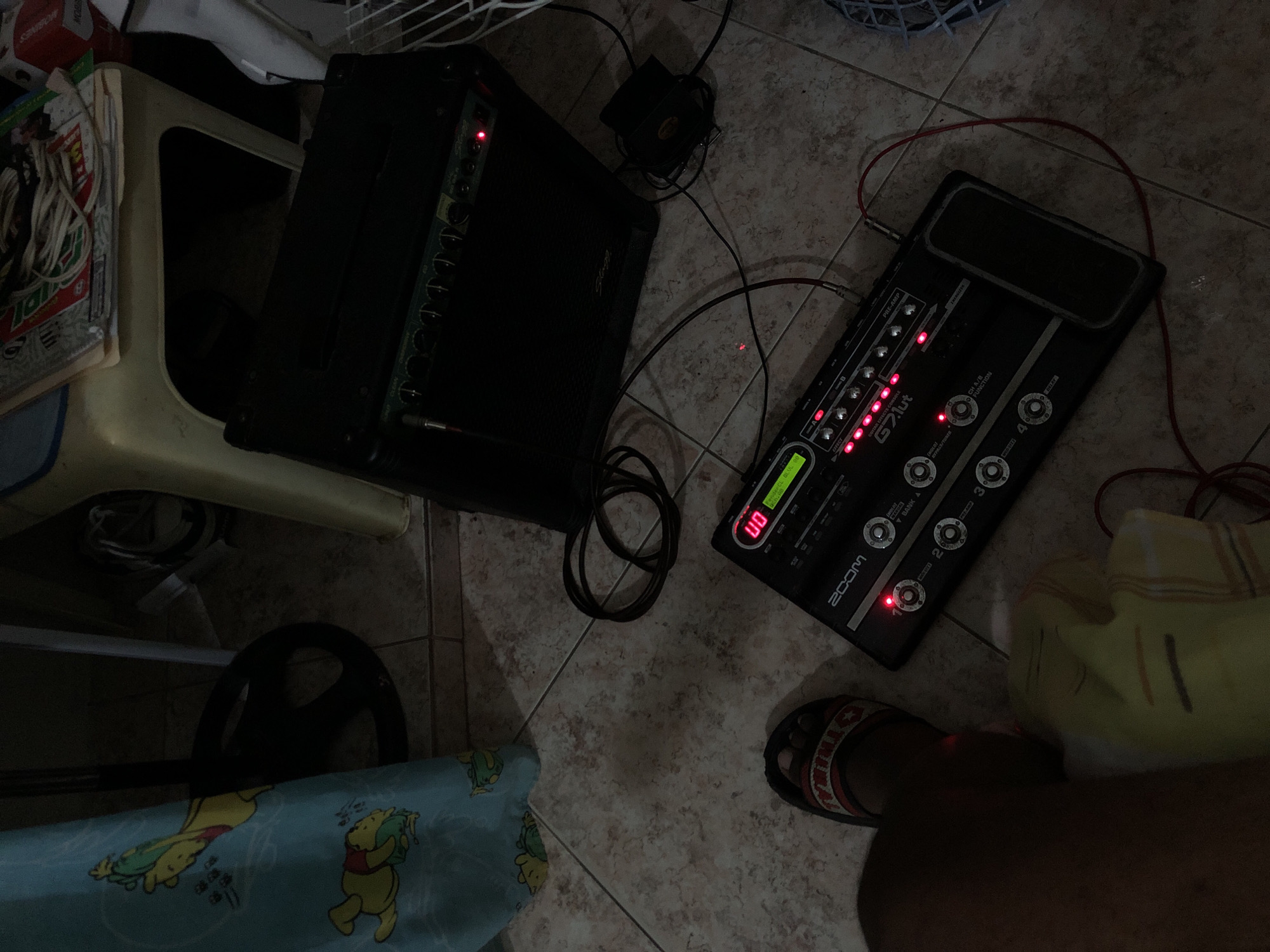 Here's a forensic description of the image:
**Scene:** The image depicts a section of a tiled floor. A small amplifier is located next to a white plastic chair stacked with books and other paper materials. Adjacent to the amplifier is a black multi-effects guitar pedal, model "Zoom G7.lut". Various cables connect the pedal and amplifier, with some slack coiled on the floor. A power adapter is visible between the two pieces of equipment. A foot wearing a sandal is partly visible next to the pedal. Part of a Winne the Pooh themed child’s bed sheet appears to be at the bottom left. A leg and part of a cushion is visible bottom right.
**Environment:** The environment appears to be an indoor setting, possibly a bedroom or practice space. The lighting is dim.
**Location:** There is no evidence to suggest this image was taken in Altagracia, Venezuela.