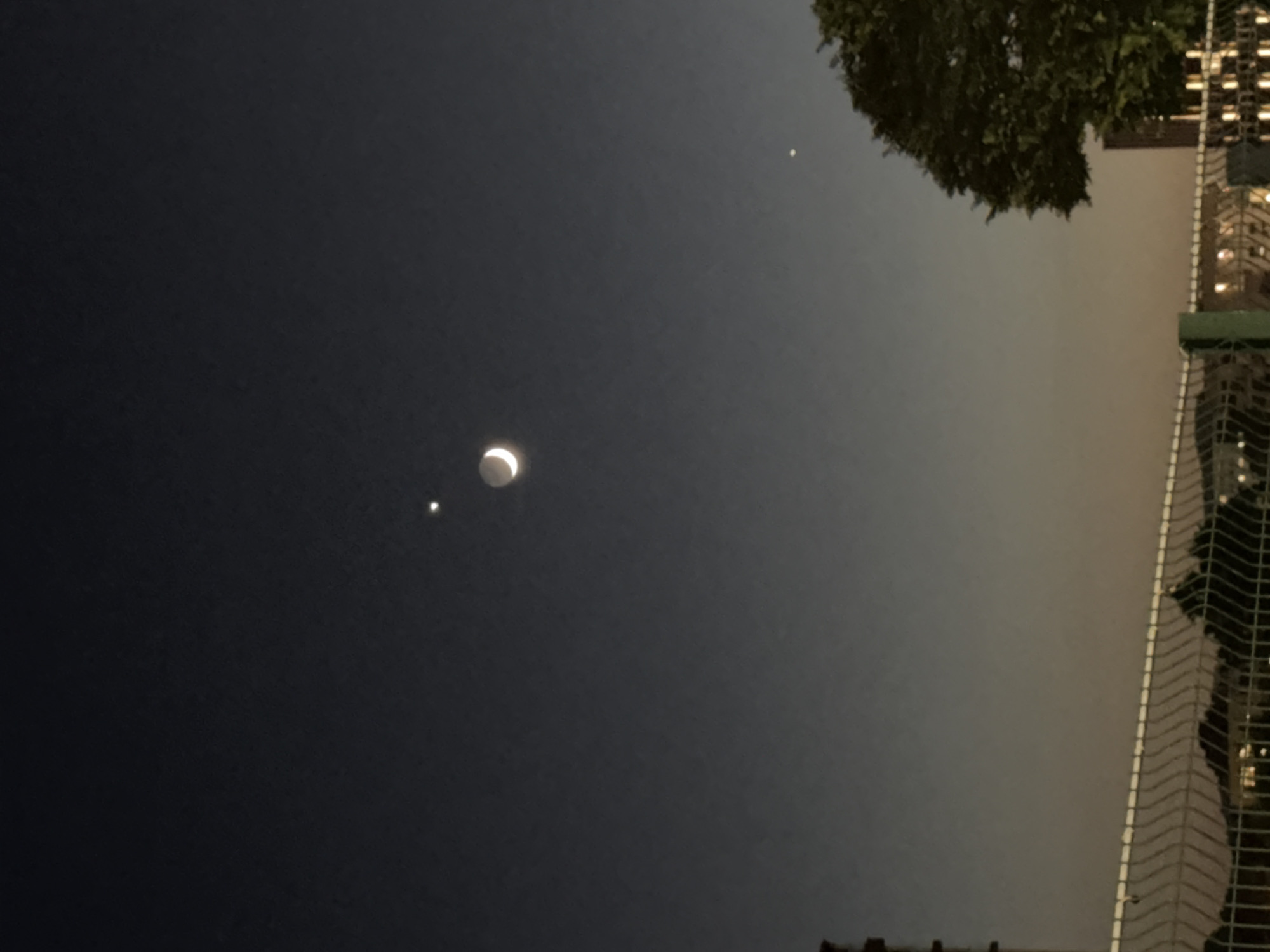 The image shows a nighttime sky over Ilidža, Bosnia and Herzegovina. A crescent moon and two bright stars or planets are visible. In the foreground, a chain-link fence runs horizontally across the bottom of the frame. Beyond the fence, there are some trees and buildings. The sky is dark, with a gradient of lighter color near the horizon. The overall impression is one of a peaceful, clear night.