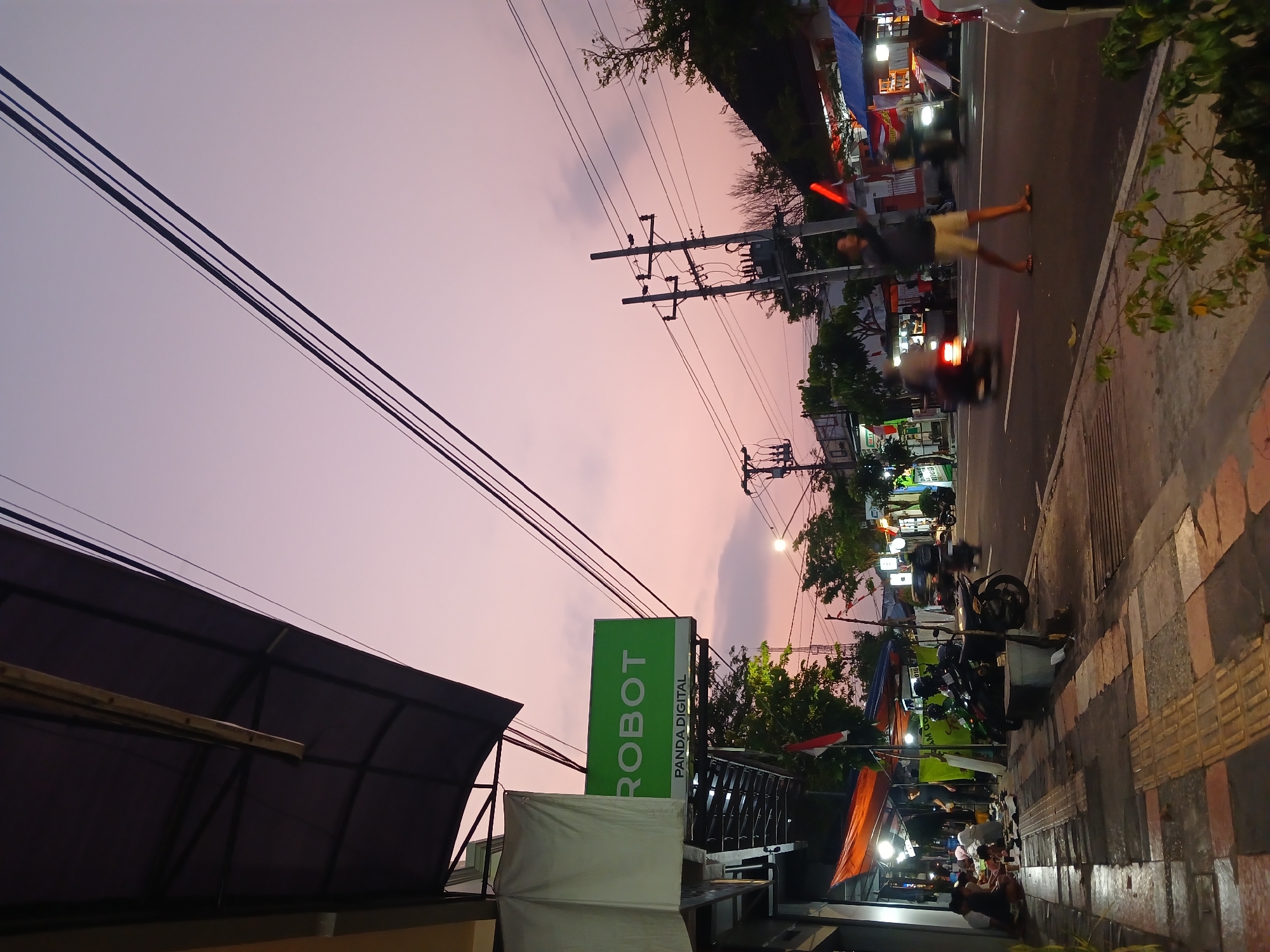 The image depicts a street scene in Temanggung, Indonesia, during the twilight hours. The sky displays a soft gradient of pink and purple hues, with several power lines crisscrossing the frame. On the left side of the street, a shop with an awning displays a green sign that reads "ROBOT PANDA DIGITAL". In front of the shop, several people are seated on the sidewalk, appearing to be socializing or eating. Various stalls and shops line the street, some with colorful awnings and lights, suggesting a commercial area or market. A person is walking across the street holding two red illuminated objects, possibly for signaling. Motorcycles are visible on the road, with some moving and others parked. The overall atmosphere is that of a bustling street at dusk, with a mix of commercial activity and pedestrian presence.