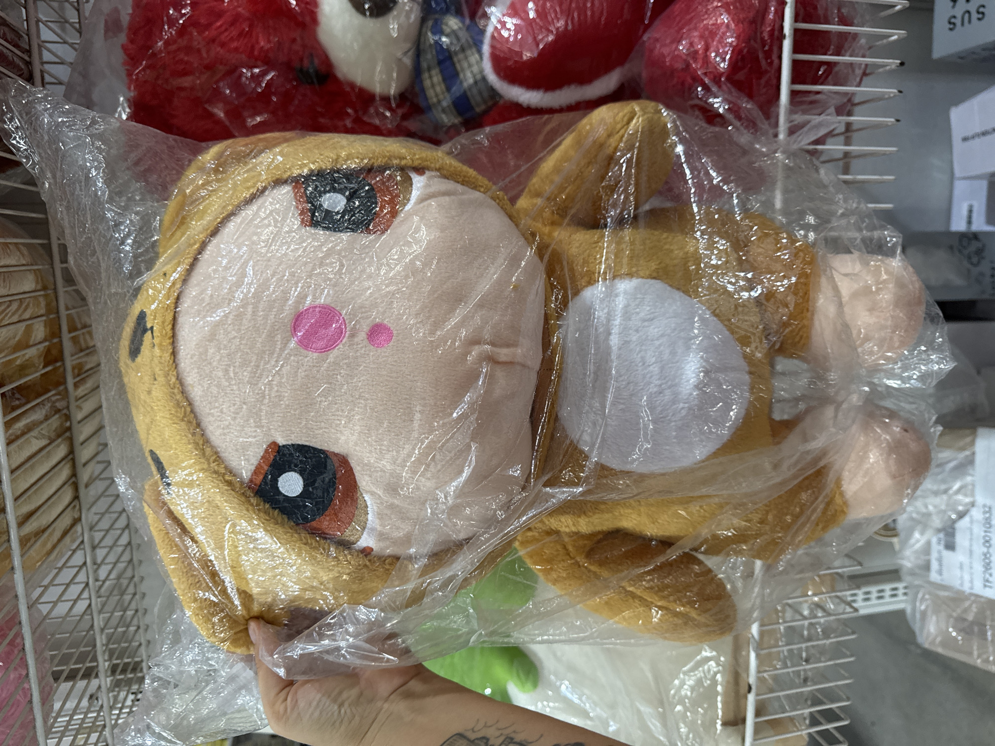 A slightly high-angle, indoor shot shows a plush toy in a clear plastic bag being held by a person with a tattoo on their forearm. The toy, predominantly tan and white, has a cartoonish face with a pink nose and large eyes. Other plush toys, mostly red, are visible in the background on a wire shelf. A printed label with "TF2605-0010632" is visible to the right of the toys. The toys are in Thong Sala, Thailand.