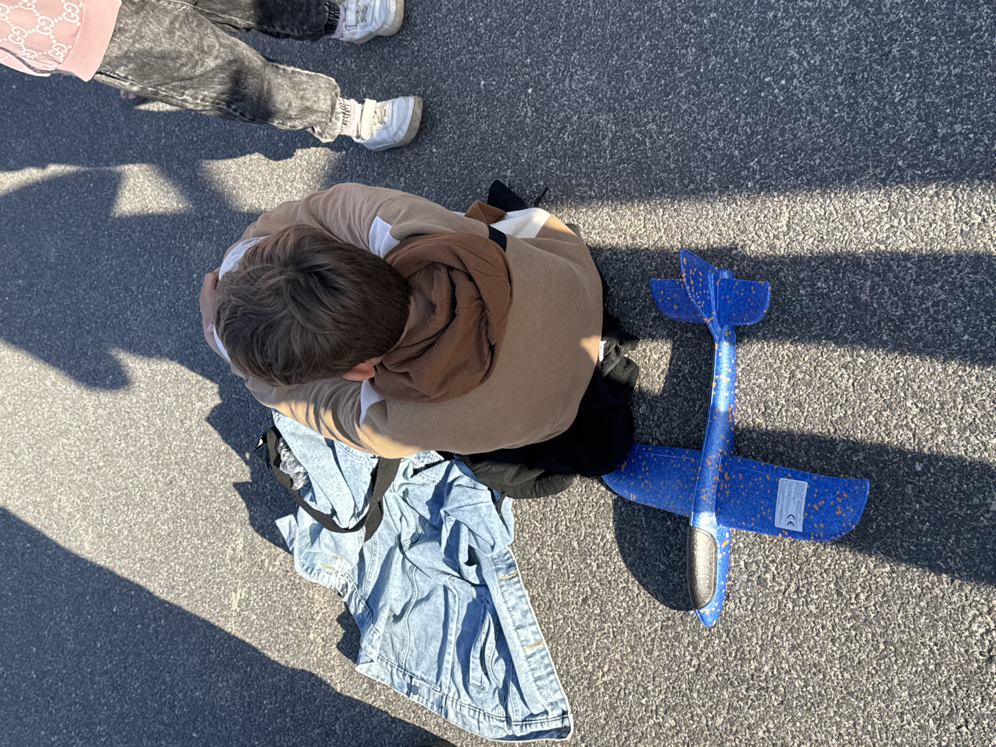 The image shows a top-down view of a young boy sitting on the ground, with his head bowed, next to a blue toy airplane. He is wearing a tan hoodie with white and black accents, and black pants. A denim jacket is laid out on the ground near him. The scene appears to be outdoors, on an asphalt surface that is partially shaded by shadows cast by what appear to be trees or buildings. To the right of the boy, another child, wearing a pink hoodie with a repeating pattern and dark, distressed jeans, stands with their legs visible. The location is identified as the city of Tallinn, Estonia.