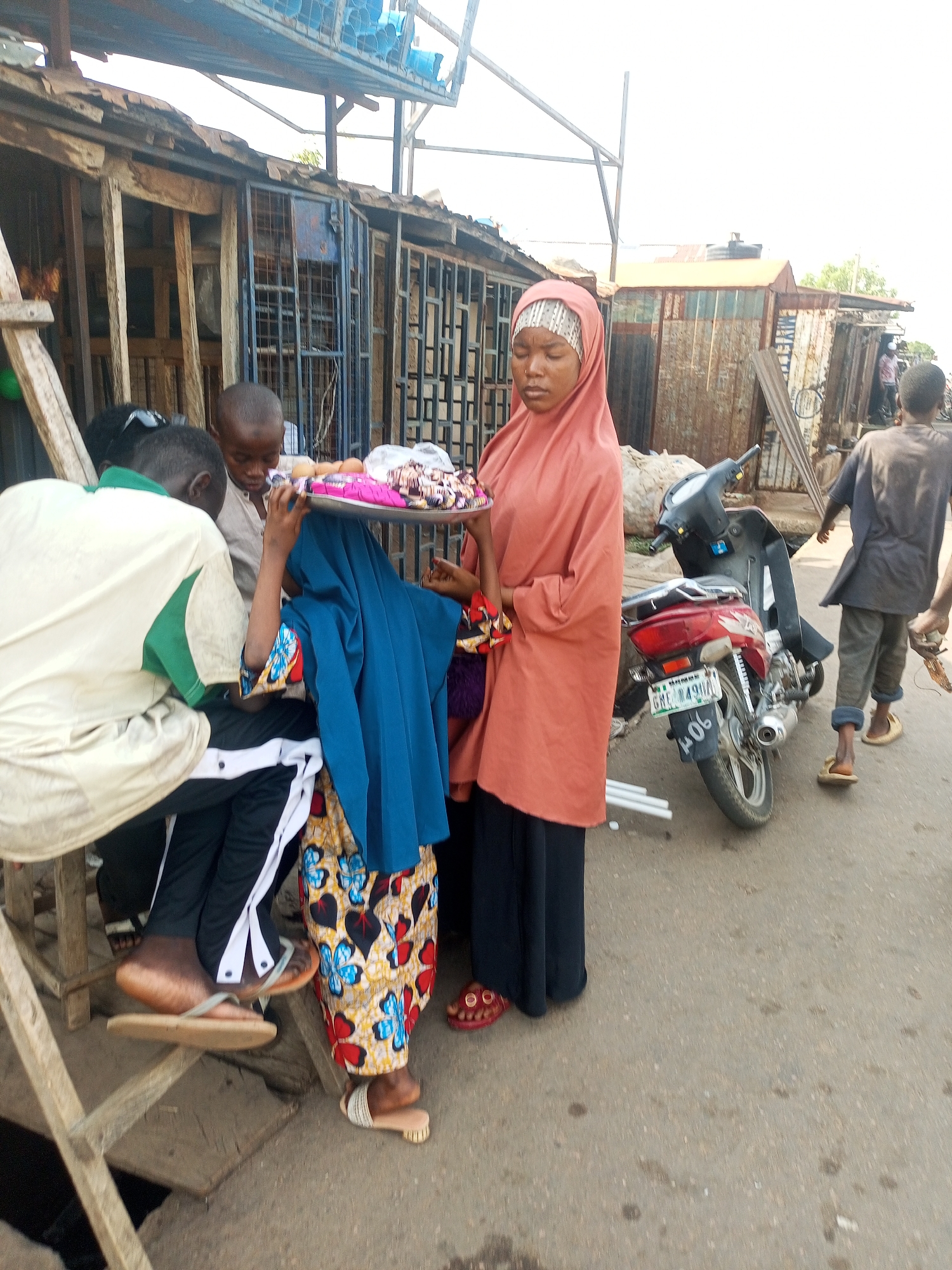 The image shows a street scene in an urban environment. A woman wearing a salmon-colored headscarf and a long black skirt stands next to a group of people who are examining goods on a tray she is holding. The tray appears to contain small items, possibly food or other wares. Several people are gathered around, one of whom is wearing a blue headscarf and a dress with a colorful butterfly pattern. Another person is sitting on a makeshift wooden structure. In the background, there is a red motorcycle parked near a corrugated metal structure, and a man walks away from the group. The location is possibly a market area or a roadside vending spot.