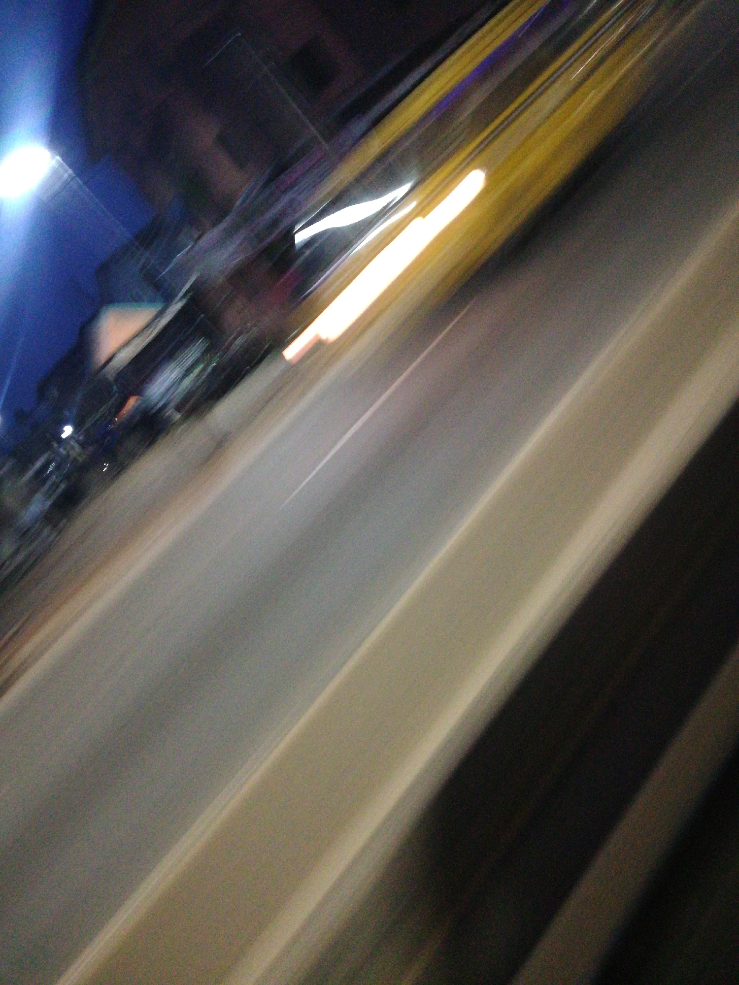 The image depicts a blurred street scene at night, likely taken from a moving vehicle. A street light is visible on the left, illuminating buildings and parked vehicles. A yellow vehicle is moving on the right, its lights blurred into streaks. The location is indicated as Lagos, Nigeria. Details regarding people, specific objects, actions, and interactions are indiscernible due to the blur. The environment is urban, suggested by the buildings and street lighting.