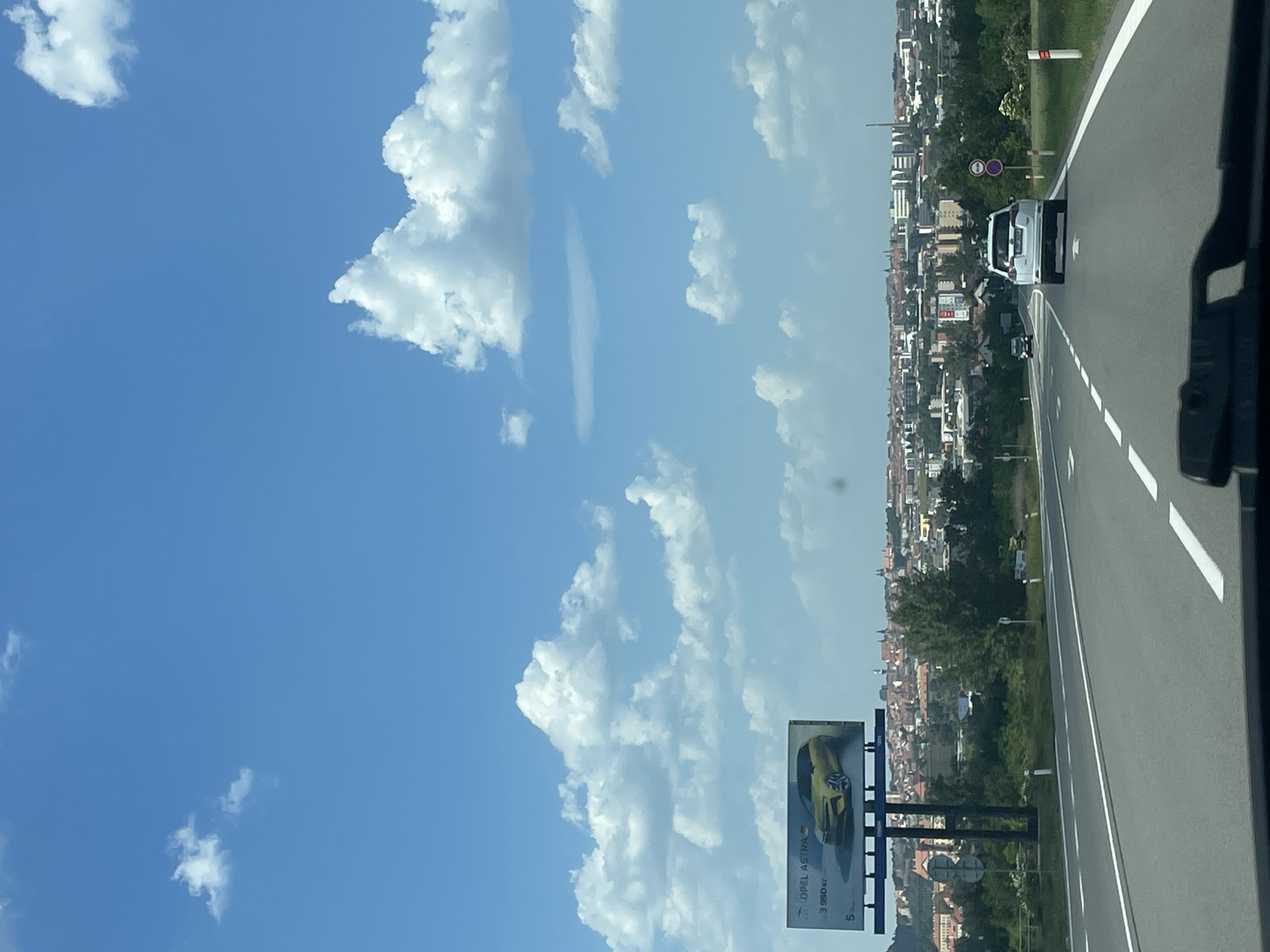 The image shows a highway on the outskirts of Znojmo, Czechia, on a sunny day. The highway is multi-lane with white line markings and appears to be relatively empty, with a white SUV in the middle lane. To the right of the highway is a view of Znojmo, with buildings and trees visible. A billboard is visible, advertising an Opel Astra car. The sky is predominantly blue with numerous fluffy white clouds. The image appears to have been taken from inside a vehicle, given the edge of the vehicle is visible in the right.