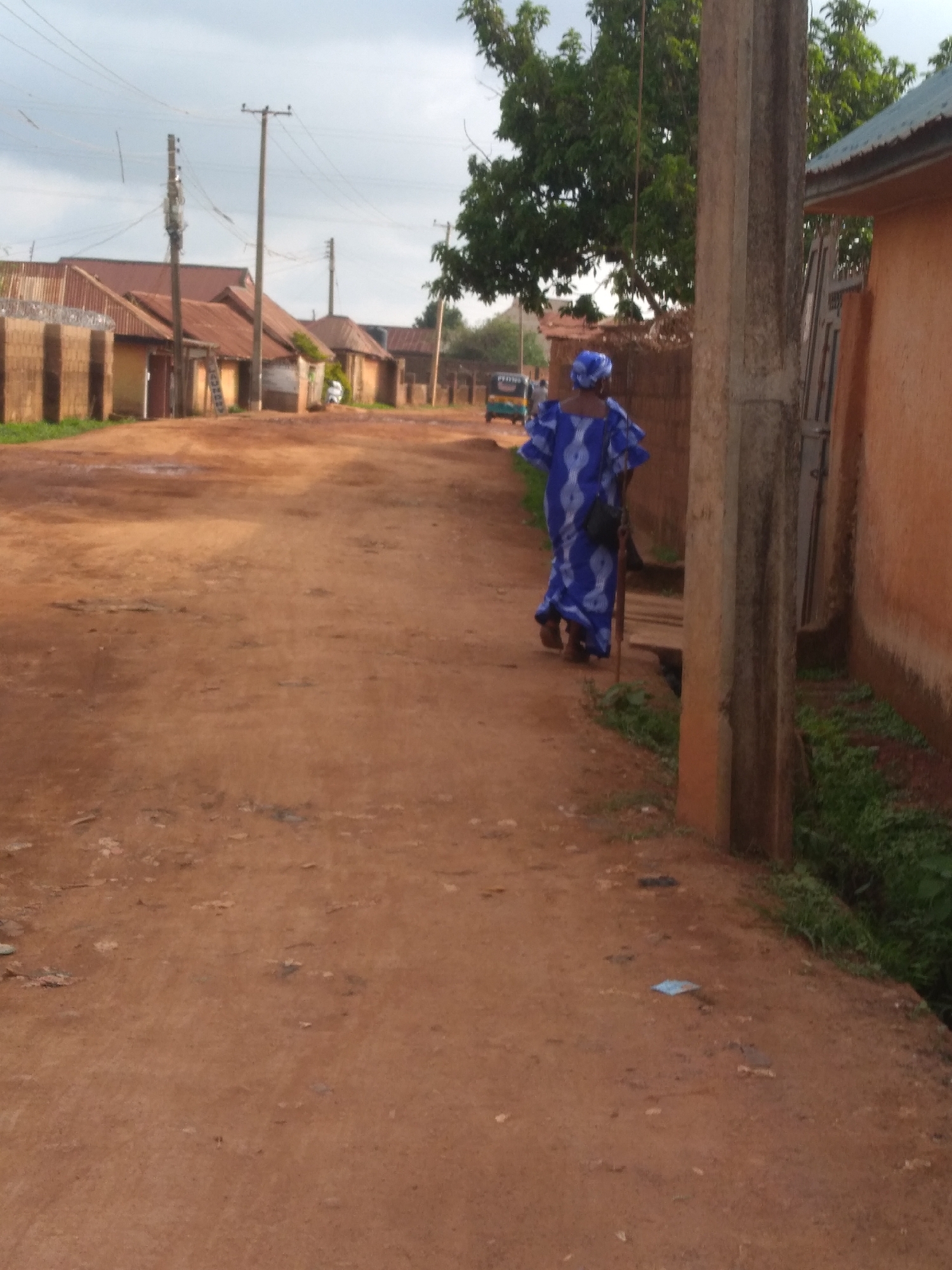 An outdoor, full-shot, eye-level, medium-angle view captures a dusty, reddish-brown dirt road in what appears to be a residential area. A woman, dressed in a vibrant blue and white patterned traditional African dress with ruffled sleeves and a matching headscarf, walks away from the camera towards the right side of the frame. She carries a black bag slung over her shoulder and uses a walking stick for support.
The road is uneven with some puddles of water, indicating recent rain. On the left side of the road, a row of single-story houses with corrugated metal roofs is visible. Some of these houses have brick walls and wooden doors. Electric poles and power lines crisscross the sky above the houses. On the right side of the road, a concrete pillar stands prominently, and beyond it, a portion of an orange-walled building with a green roof can be seen. Lush green vegetation grows along the side of the road near the building. In the background, more houses and trees line the street, and a three-wheeled vehicle is parked. The sky is overcast with light clouds. The overall impression is of a quiet, possibly rural or semi-urban setting in Nigeria, with the details suggesting a warm climate. This scene is typical of many neighborhoods in Jos, Nigeria, a city known for its dusty red earth and vibrant cultural attire.