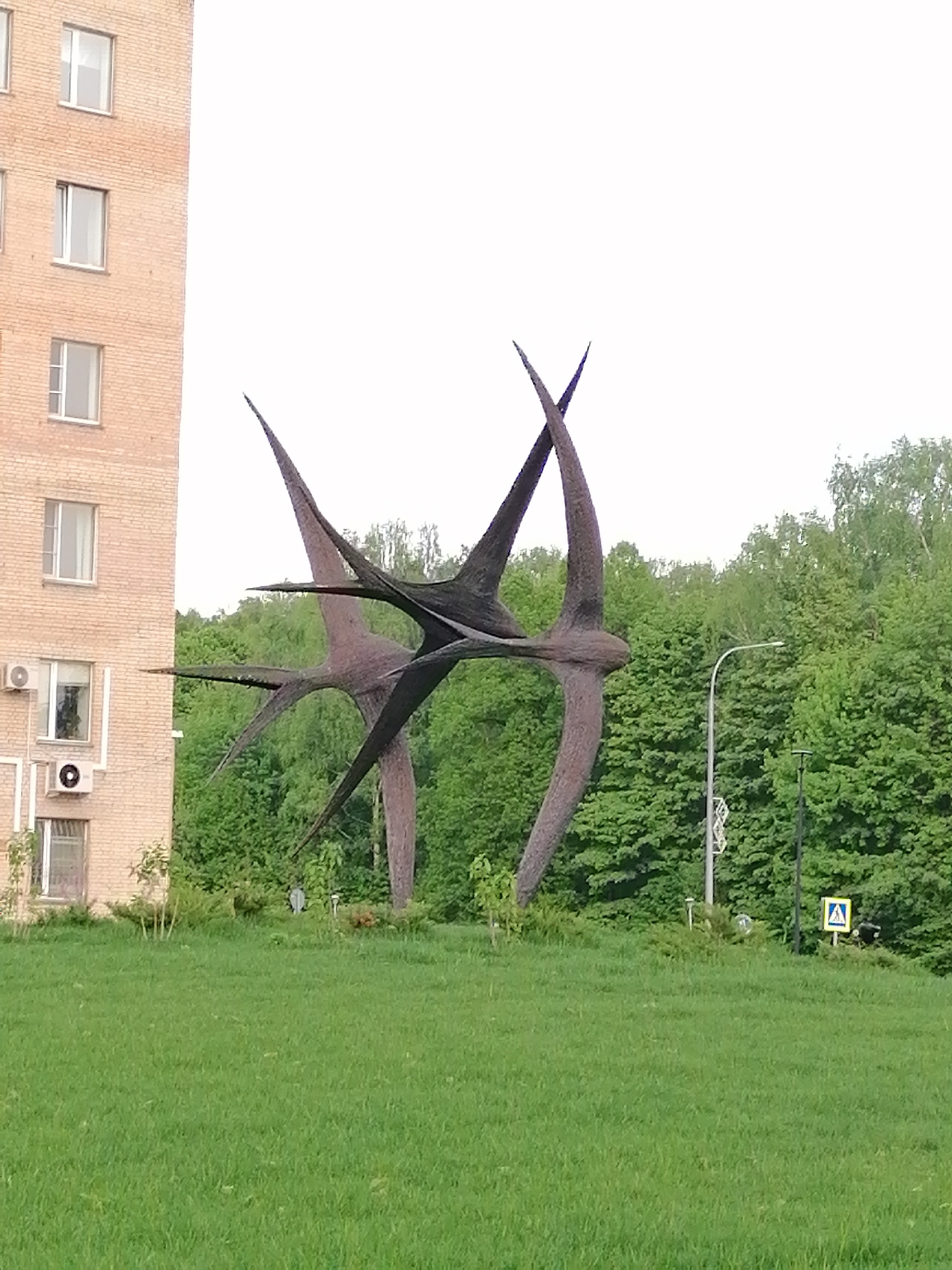 The image shows a large metal sculpture of what appears to be two abstract birds intersecting. The sculpture is situated on a grassy area in front of a brick building on the left and a wooded area with tall trees on the right. A street light is visible among the trees, and a pedestrian crossing sign stands on the edge of the grass, suggesting this is a park or green space within a city. The sky is overcast.