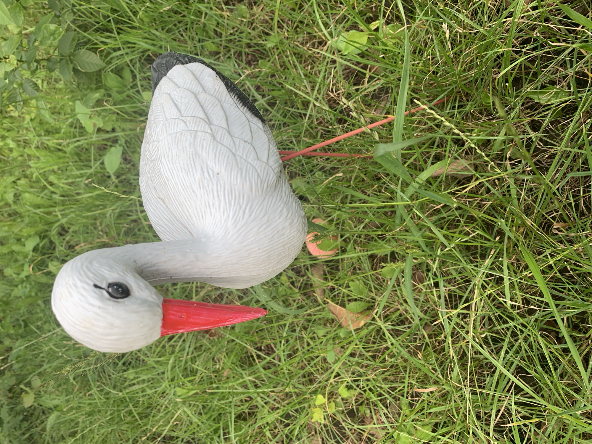 The image shows a close-up, eye-level view of a white stork figurine with a red beak and black wingtips, standing on its thin red legs amidst green grass. The figurine is positioned slightly off-center to the right. The background consists of lush green vegetation and foliage, suggesting an outdoor setting. There are no people or animals present in the image, and no recognizable landmarks indicate the specific location in Karmanovo, Russia.