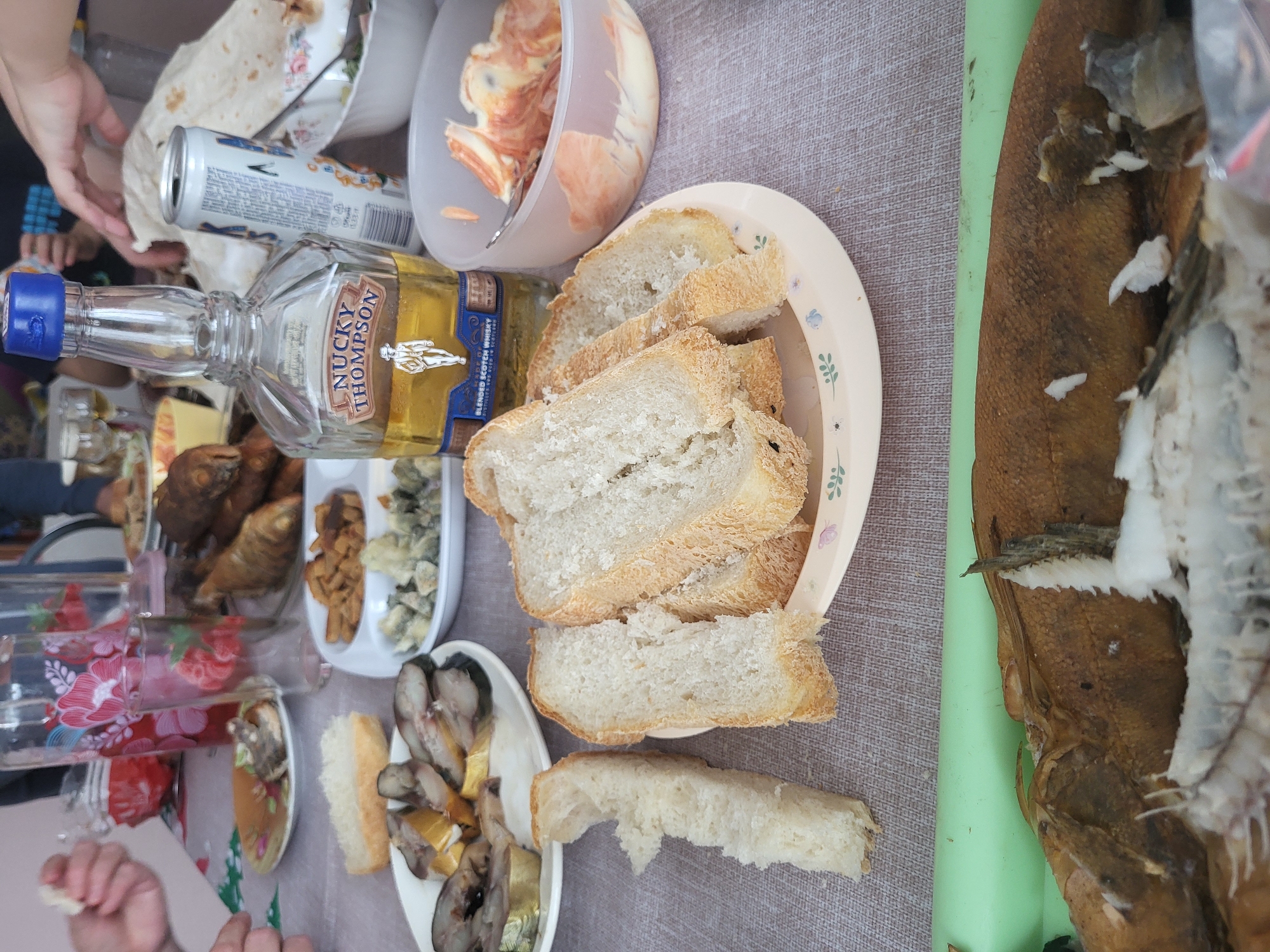 A table laden with various food items and a bottle of Nucky Thompson blended scotch whisky is visible. The table, covered with a linen tablecloth, presents a variety of dishes, including sliced bread on a plate, smoked fish, canned goods, and various salads. A half-eaten flatfish with visible bones and skin occupies a prominent space on the right side of the frame. The setting suggests an informal gathering or meal. The location, Зипуново, Russia, is noted. The scene appears to be indoors. There are hands of people that can be seen around the table, which suggests that there are people involved.
