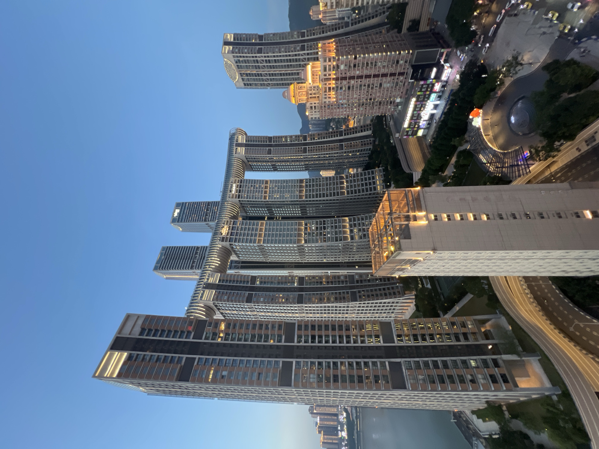 The image shows a nighttime cityscape in Yuzhong District, China, dominated by the distinctive "Crystal" or "Gateway" building complex. This architectural marvel features multiple skyscrapers connected by a horizontal, curved structure resembling a tunnel or bridge, with integrated lighting. Several other tall, modern buildings are visible surrounding the main complex, some with illuminated windows and facades. A river or body of water is present on the left side of the frame, reflecting the city lights. A winding road with streetlights and moving vehicles is seen in the foreground and midground, suggesting urban traffic. Green spaces, possibly parks or landscaped areas, are interspersed between the buildings. The sky is a deep blue, indicating either twilight or a clear night. The scene is bustling with urban activity and architectural innovation.