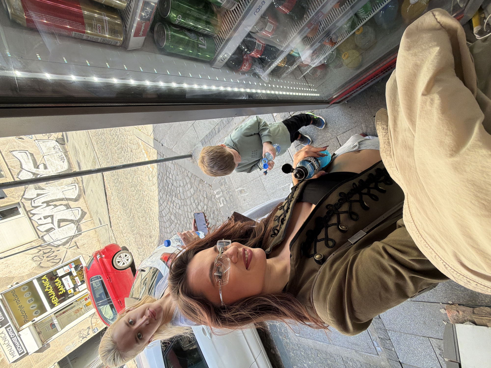 The image captures a candid, high-angle outdoor scene in an urban setting, likely Belgrade, Serbia, on a bright, sunny day. Three individuals are prominently featured. In the foreground, a woman with long, dark hair, wearing glasses and a military-style olive green jacket, looks directly at the camera with a soft smile, holding a blue Powerade bottle. Next to her, partially visible, is another woman with blonde hair, also looking towards the camera.
Further back, a young child with light hair, wearing a green long-sleeved shirt, stands facing a large, glass-fronted commercial refrigerator, holding a clear plastic bottle. The refrigerator is well-stocked with various canned and bottled beverages, including red cans marked "6600", green cans of "Зајечарско" (Zaječarsko) beer priced "120", Coca-Cola products, and "rosa" bottled water. LED lights illuminate the shelves within the fridge.
The background reveals an urban street environment with cobblestone paving and graffiti on a building wall, including the text "FAKE 223". A red car is parked on the street. A kiosk or building to the left displays multiple signs, clearly visible are "MENJAČNICA" (Exchange Office), with "EXCHANGE OFFICE" written below it, "Plaćanje računa" (Bill payment), and "лото" (Lotto). The overall impression is a casual stop during a daytime outing.